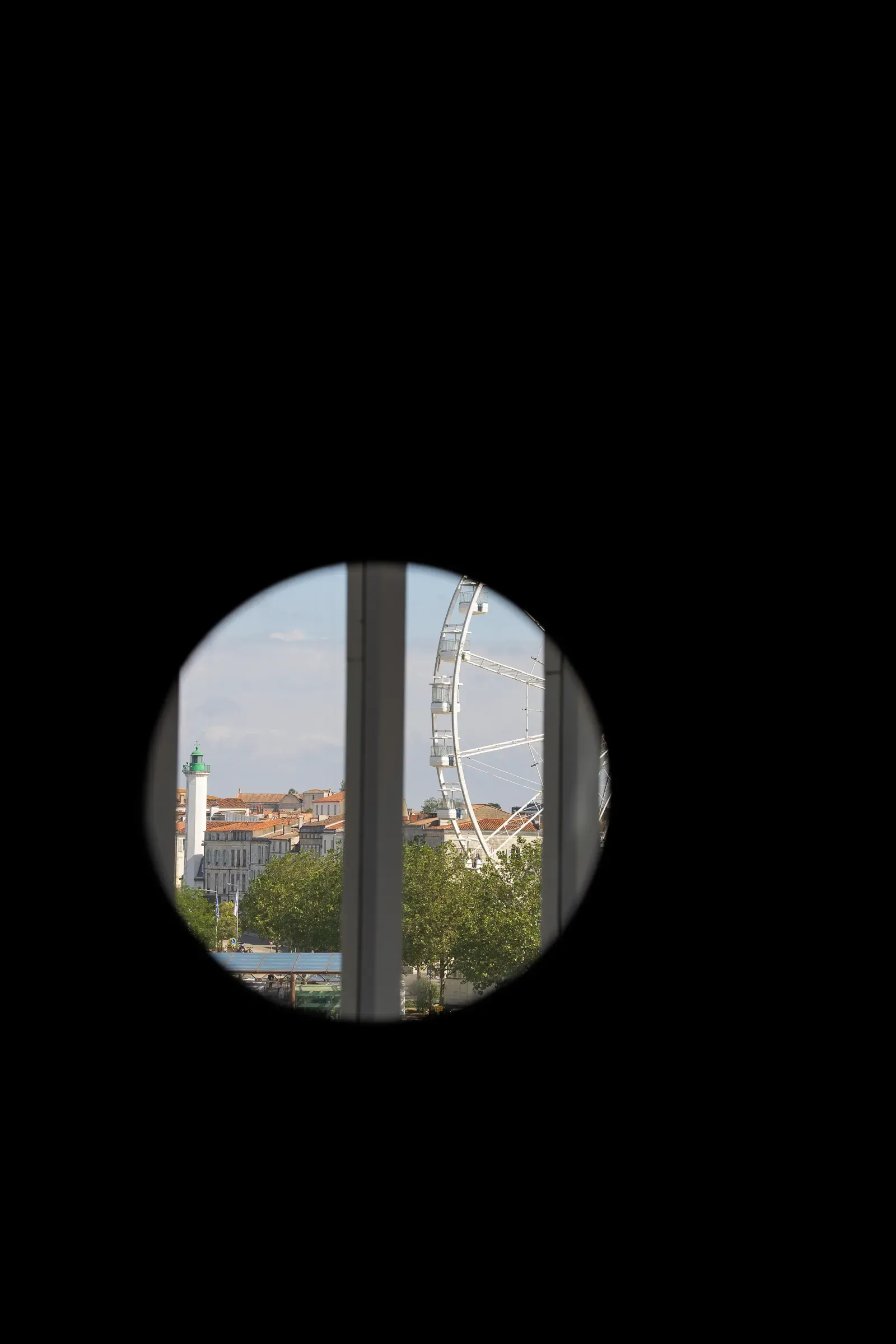 La Rochelle's lighthouse and Ferris wheel framed through a circular porthole opening, dark surround, orange rooftops and trees visible in the round window