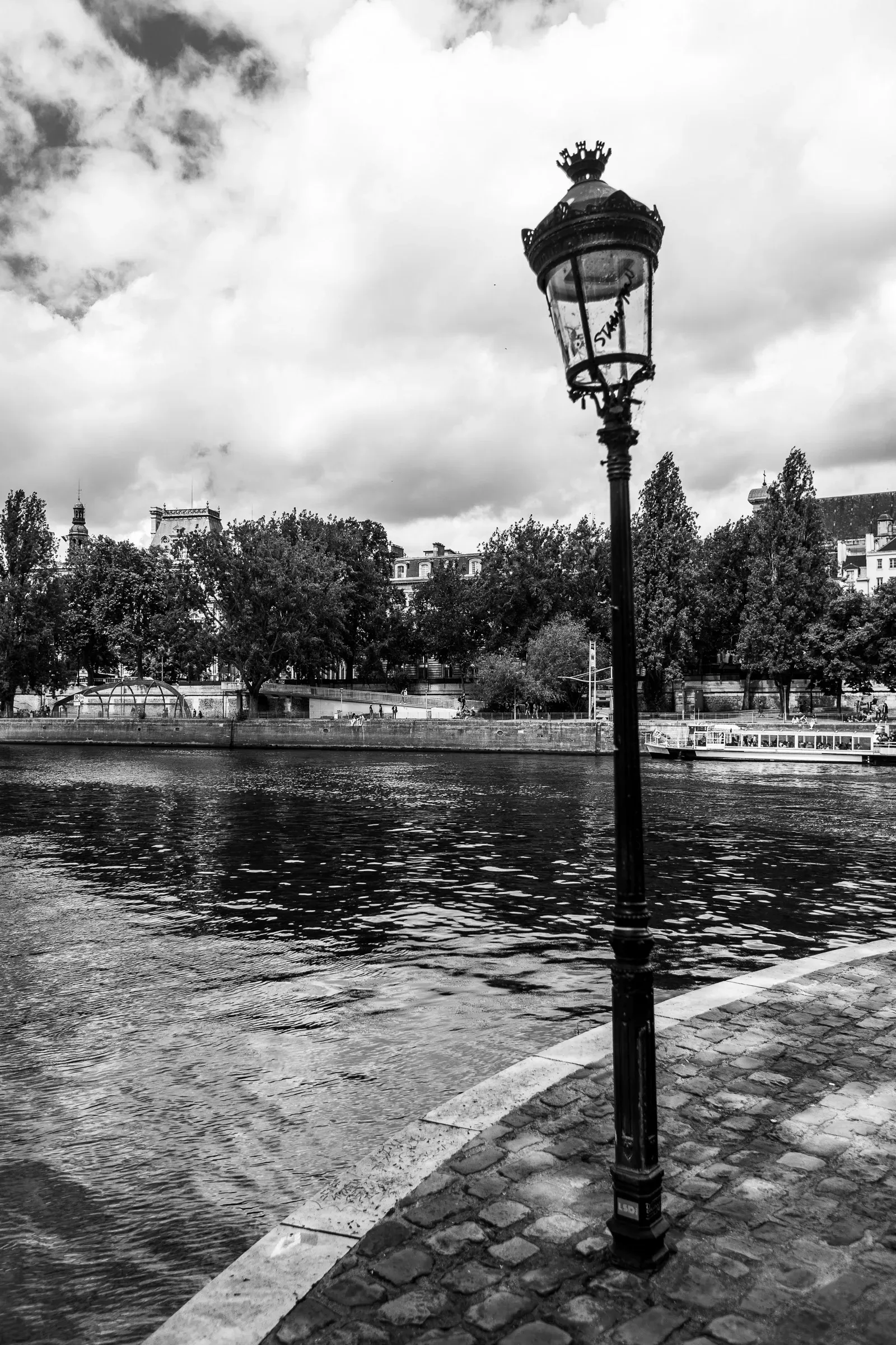 Ornate Parisian street lamp standing on a cobblestone quai beside the Seine, trees and buildings across the river in soft focus