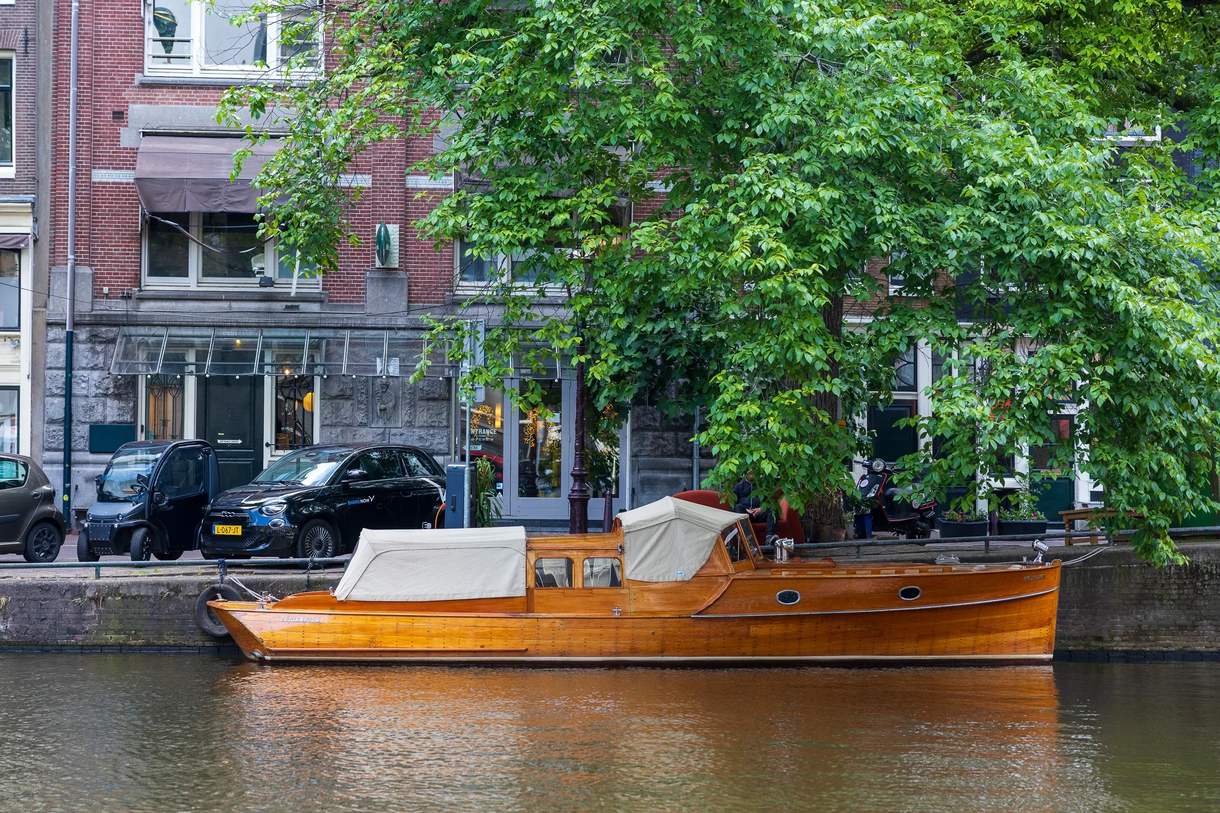 Varnished wooden motorboat moored on a canal, red brick facade with large shop windows behind