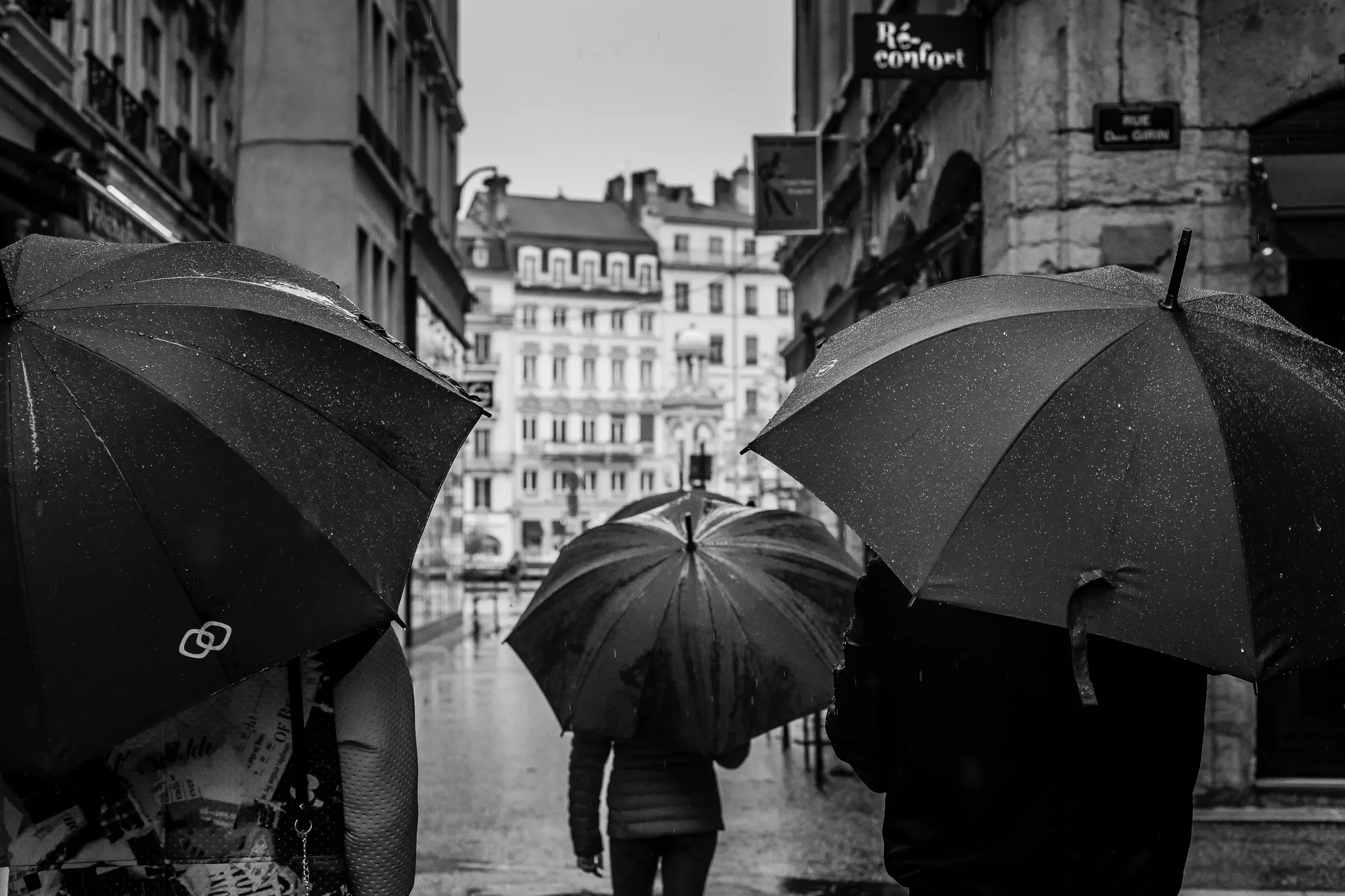 Three open umbrellas crowd a rain-soaked street, buildings rising in the background