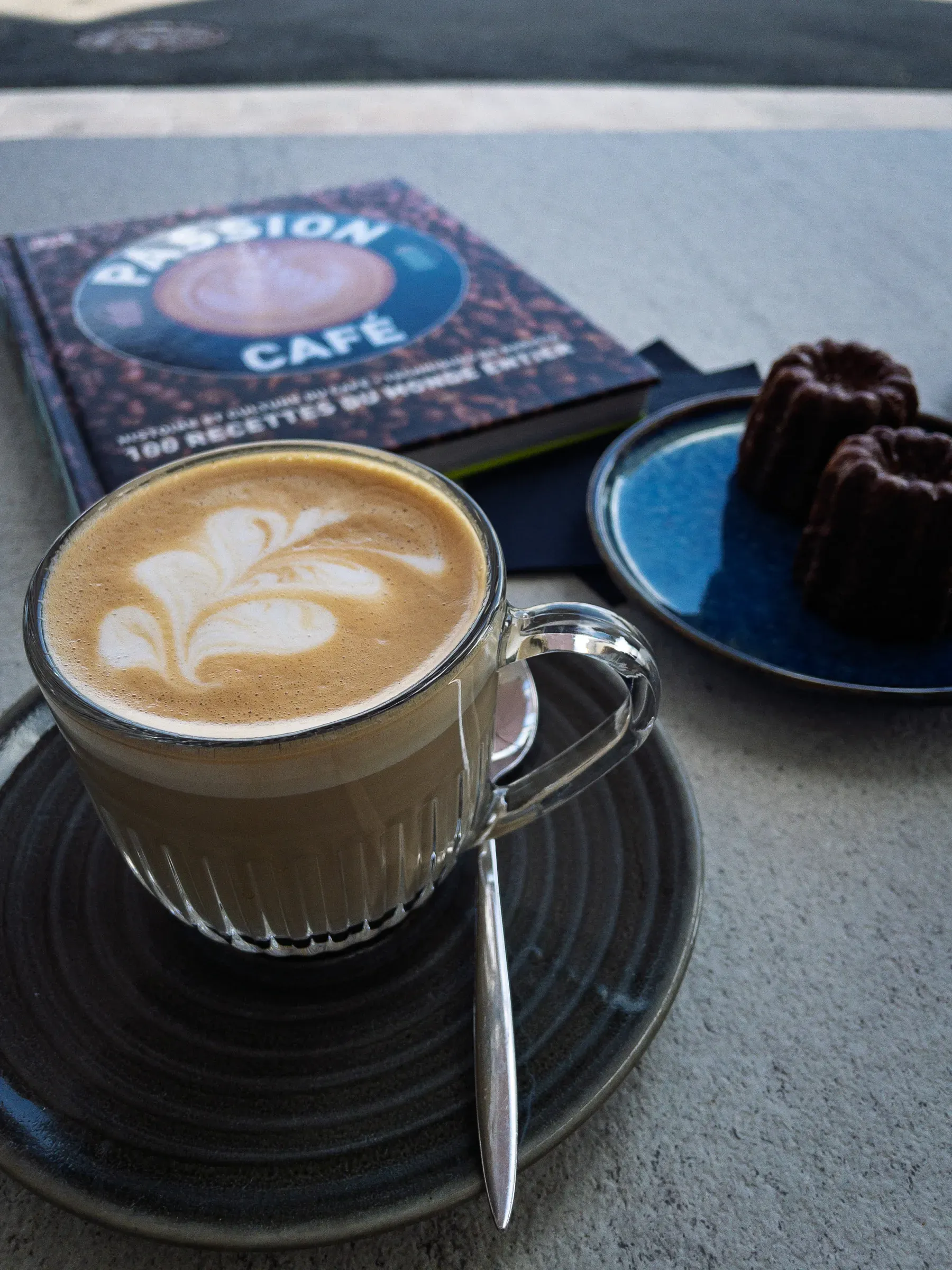 Flat white with latte art in a glass cup on a dark ceramic saucer, a canelé on a blue plate beside it, a coffee book blurred in the background