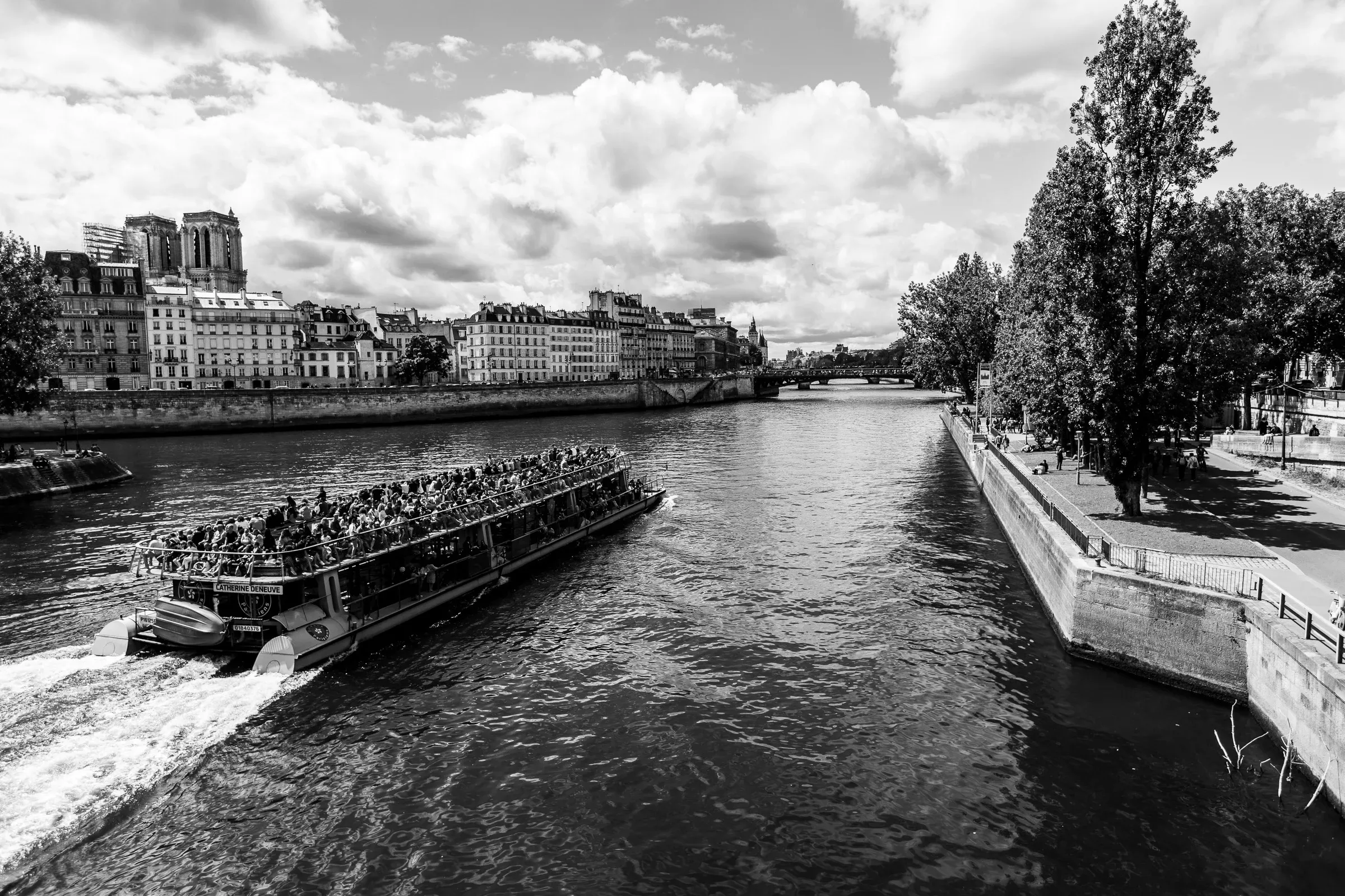 A packed bateau-mouche tour boat churning through the Seine, Notre-Dame cathedral visible on the Île de la Cité in the far left background