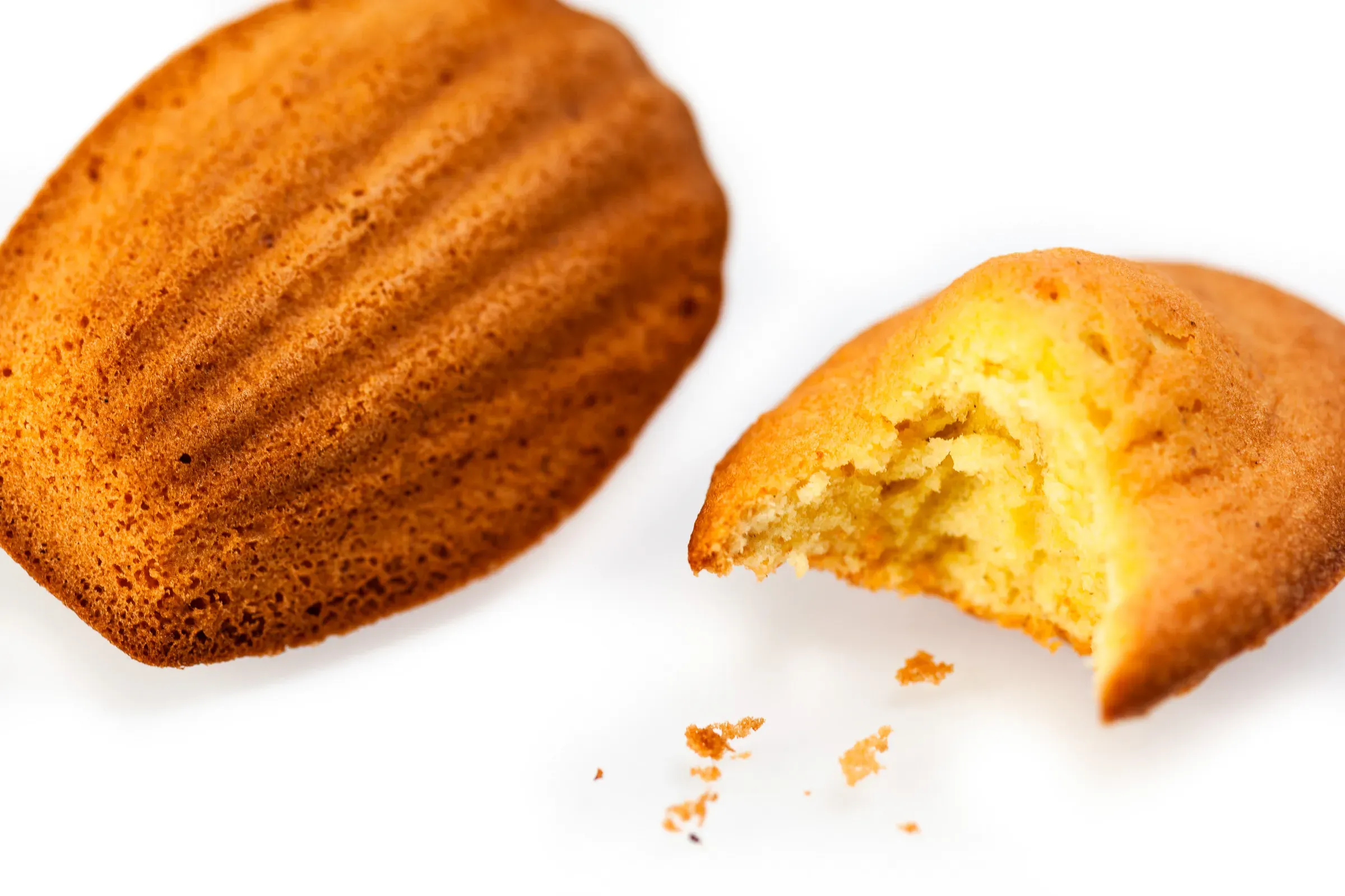 Two madeleines on a white surface — one intact shell-side up, one broken open to show the tender golden crumb inside, a few crumbs scattered