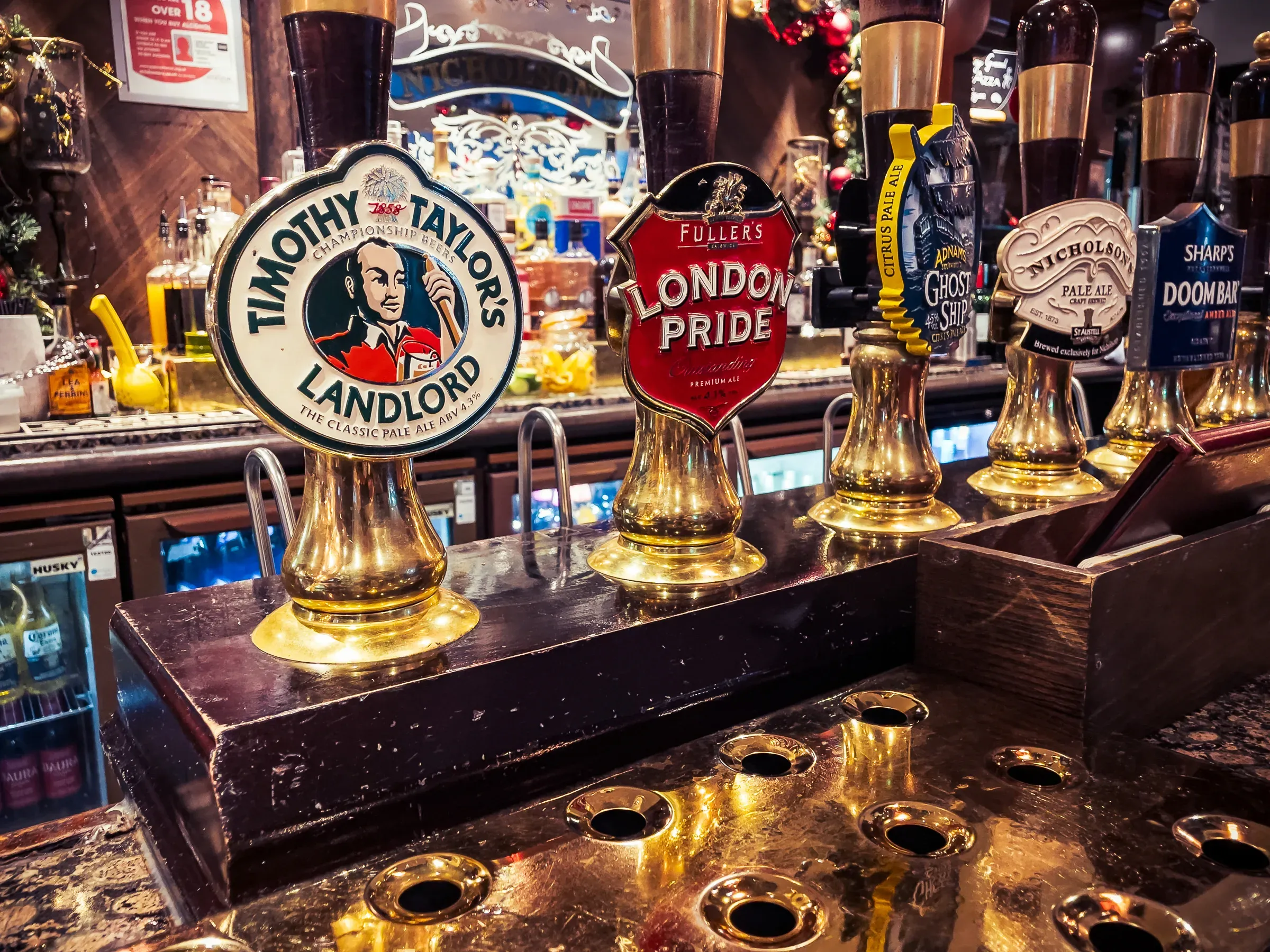 Pub beer tap handles — Timothy Taylor's Landlord, Fuller's London Pride, Ghost Ship, Doom Bar — polished brass in warm pub light