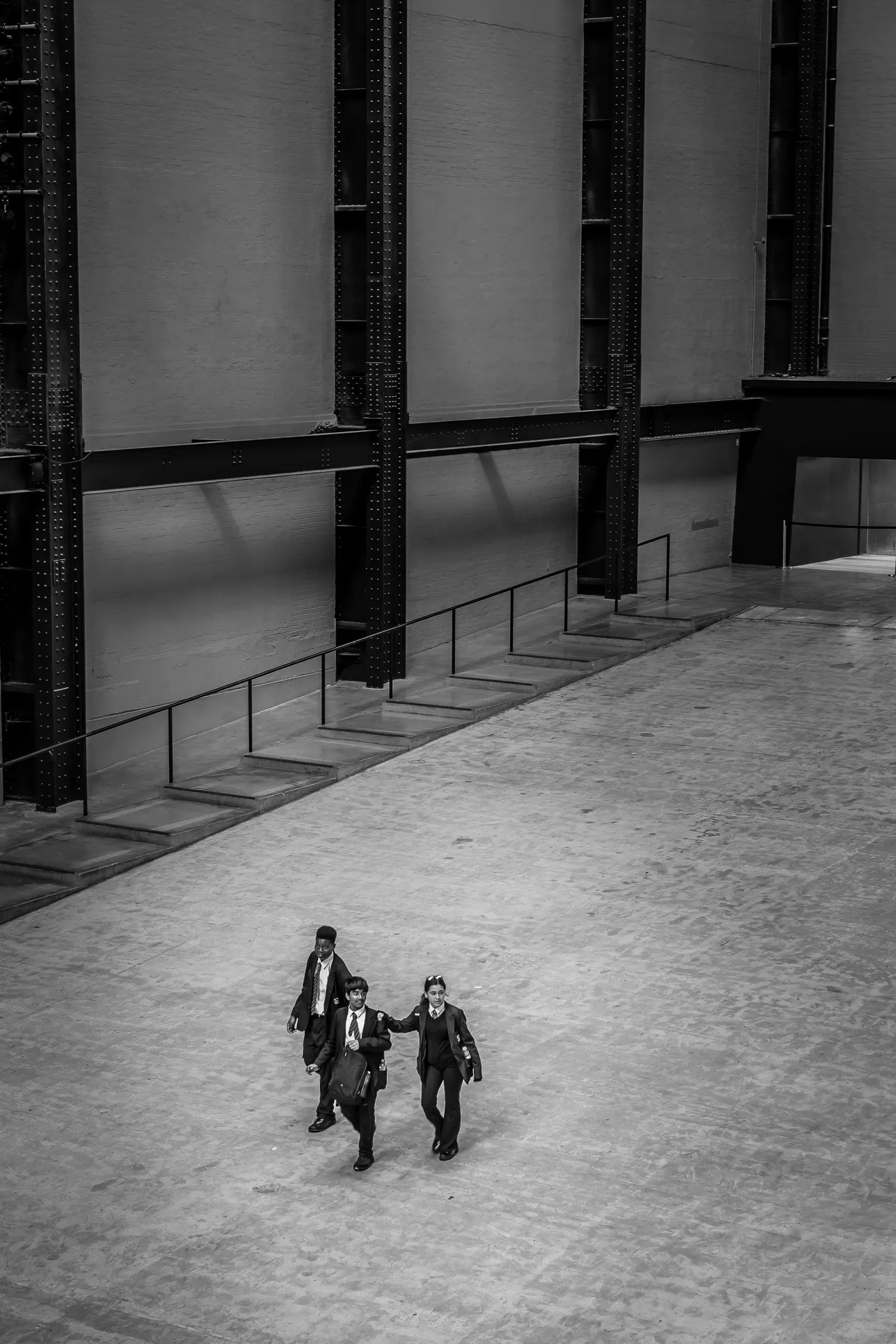Three teenagers in school uniform cross a vast concrete hall, dwarfed by rows of steel columns