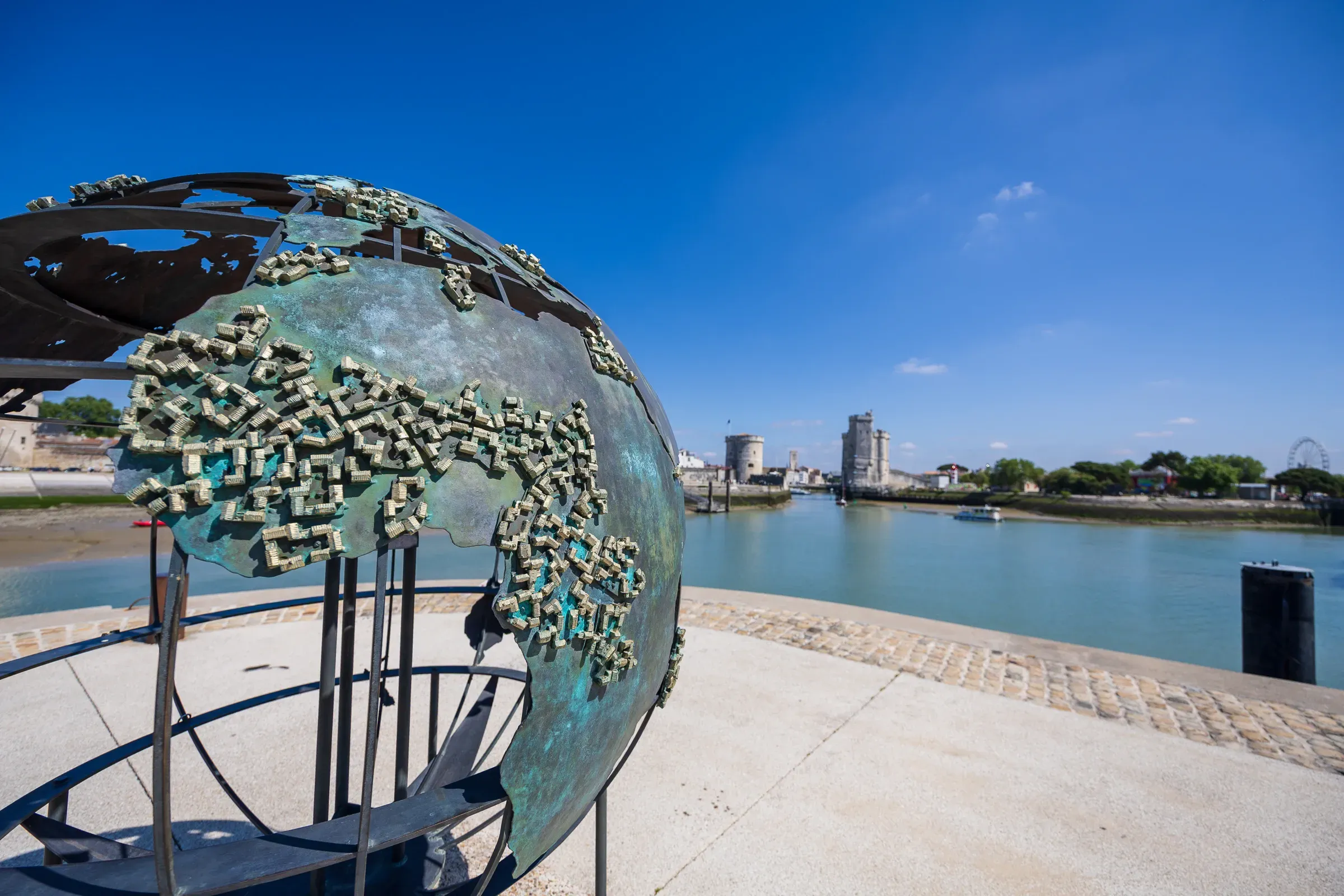 Bronze globe sculpture covered in small figures on La Rochelle's waterfront, Tour Saint-Nicolas and a second tower visible behind across the water, Ferris wheel to the far right, deep blue sky