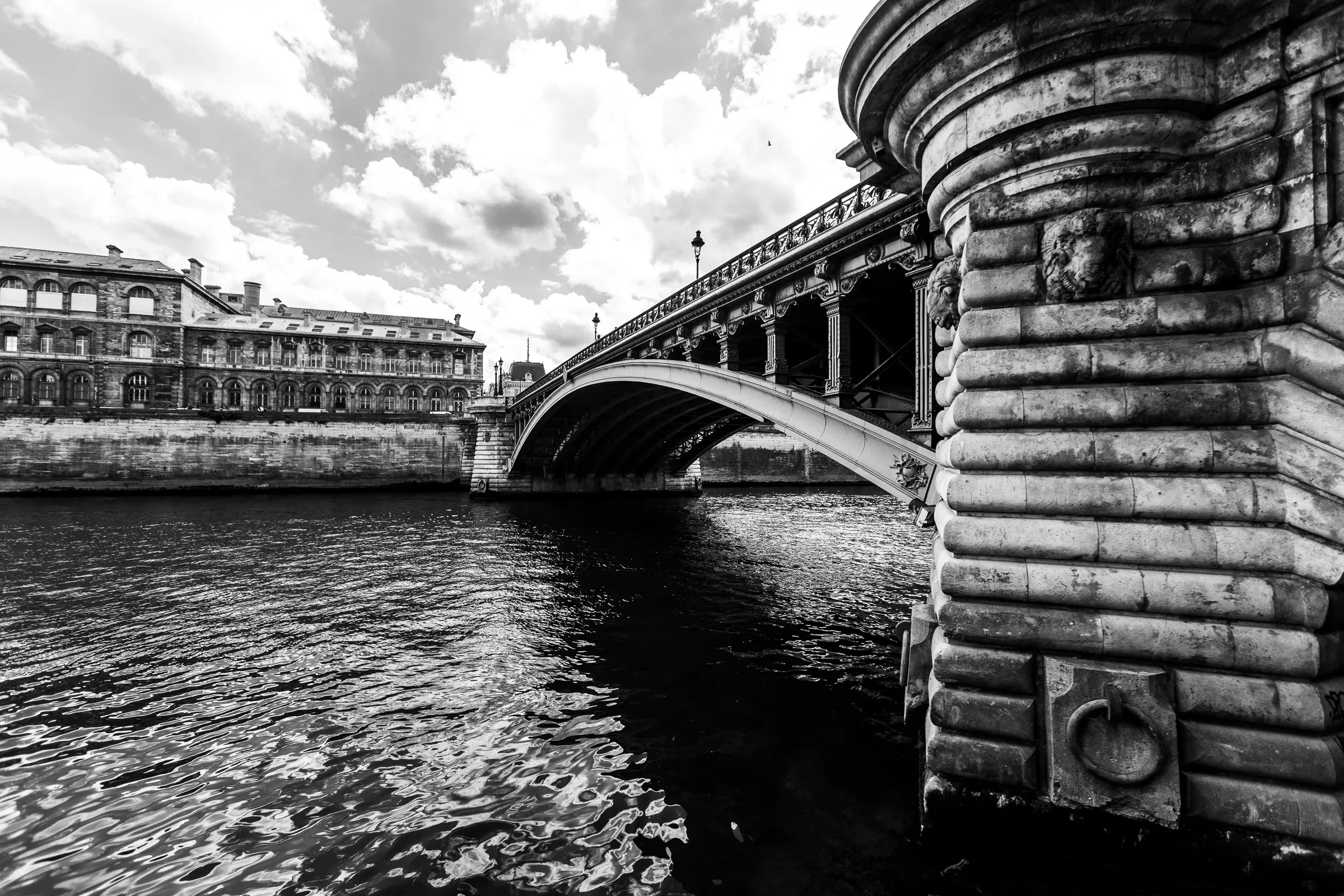 Stone arch of a Paris bridge framing the dark river below, the elaborate carved pillar close in the right foreground, a Haussmann facade behind