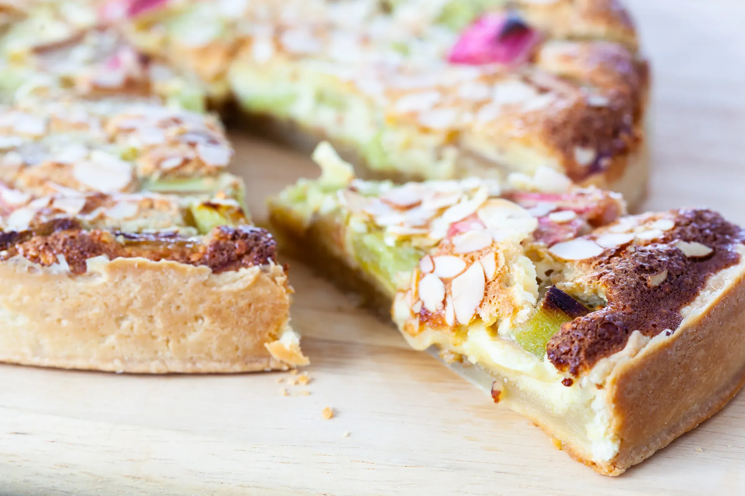 Rhubarb and almond cream tart on a wooden board, a slice pulled away to show the custard filling and flaked almond top