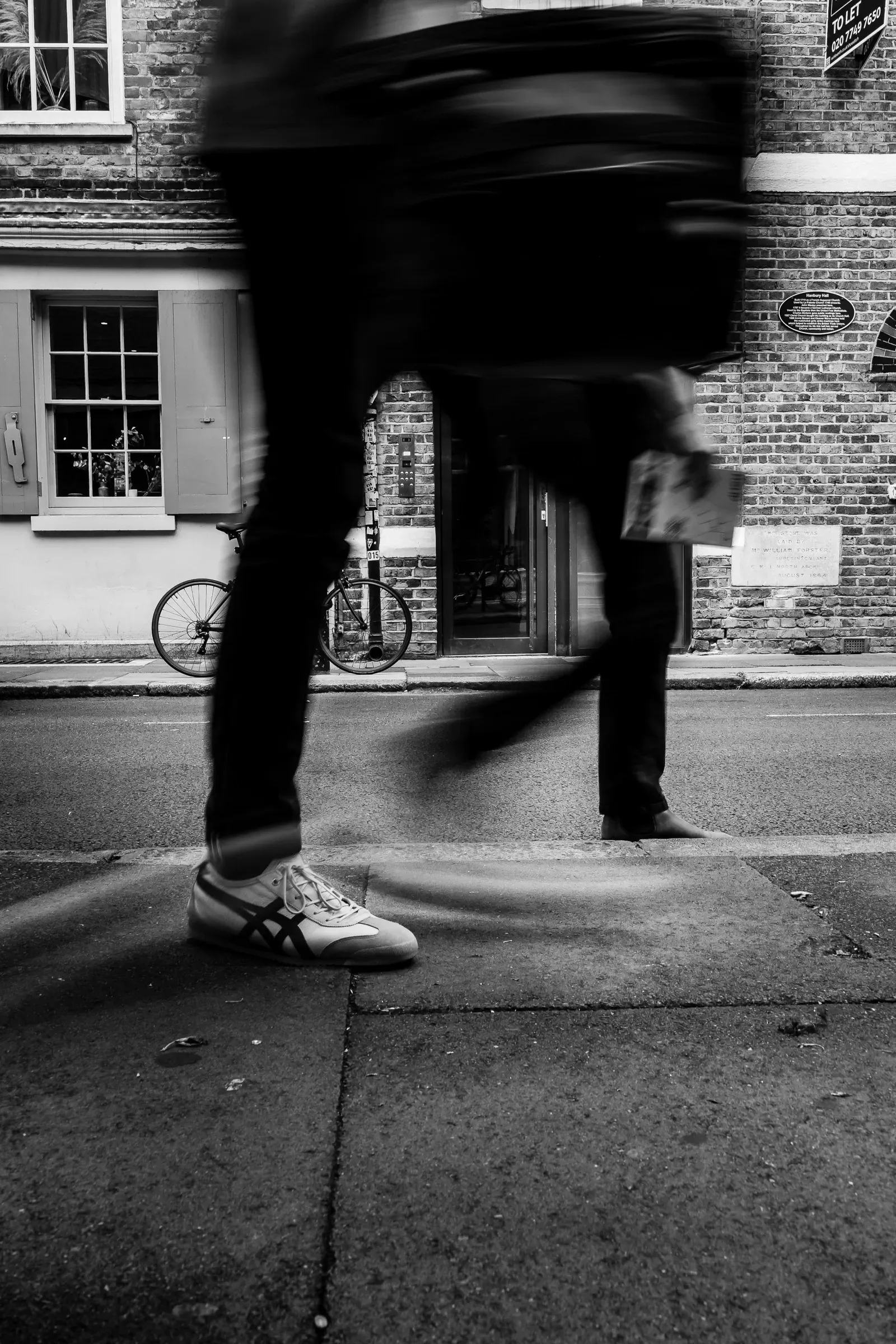 A blurred pedestrian strides past on a London street, a bicycle and the 'Hanbury Hall' plaque visible on the brick building behind