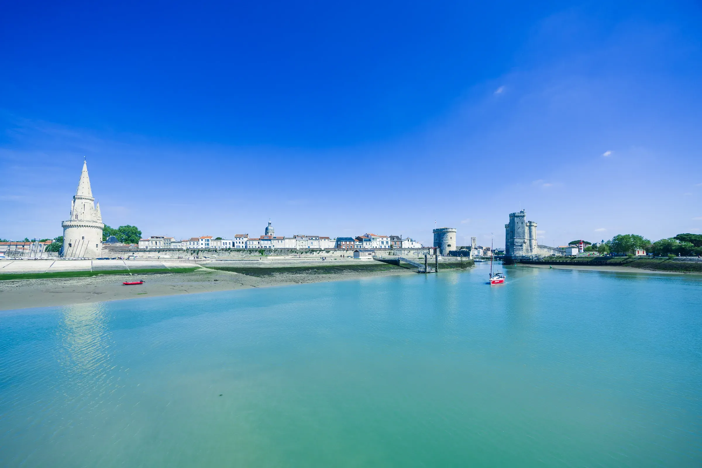 Wide view of La Rochelle's waterfront from across the harbour — the Lanterne tower on the left, the two medieval towers on the right, a red kayak and sailboat on turquoise water under a deep blue sky