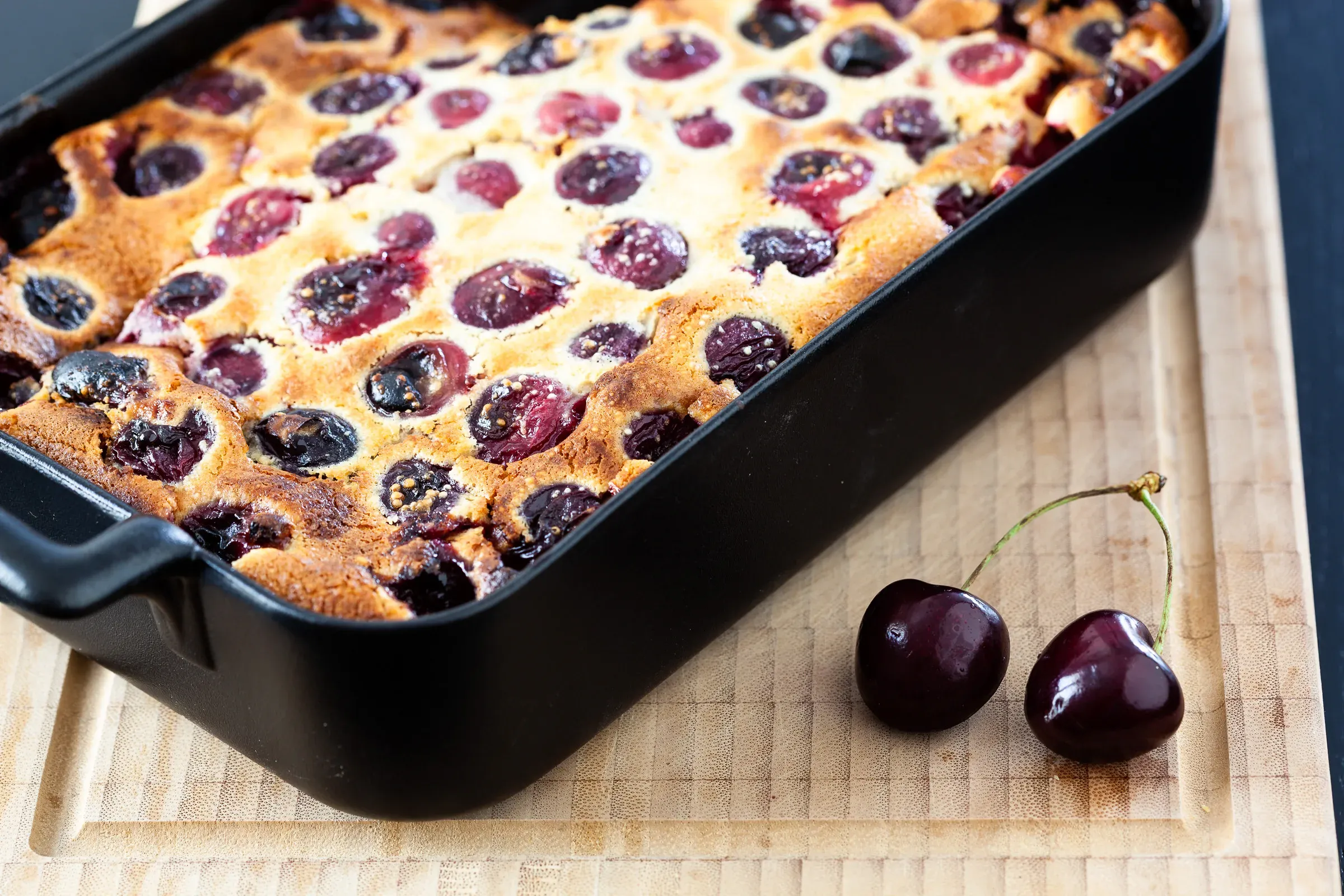 Cherry clafoutis in a black rectangular baking dish on a wooden board, whole cherries dotting the golden batter, two loose cherries beside it