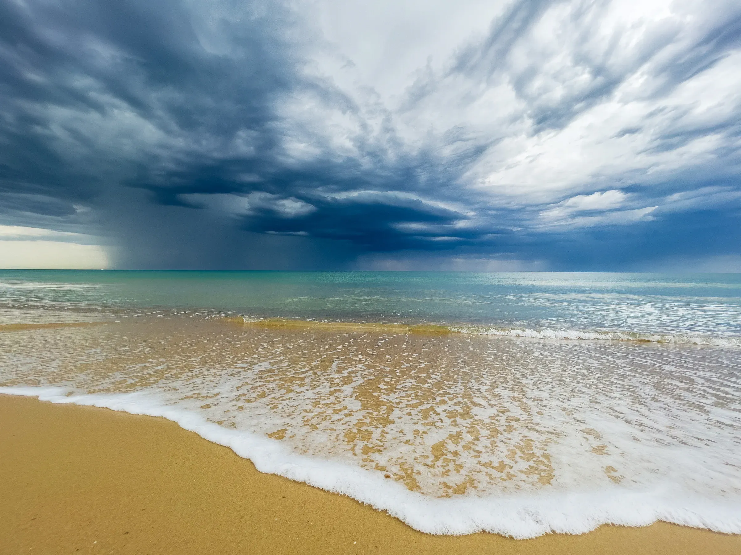 Storm front rolling over a beach, Hossegor — dark cloud mass above pale turquoise water, white foam on the shoreline