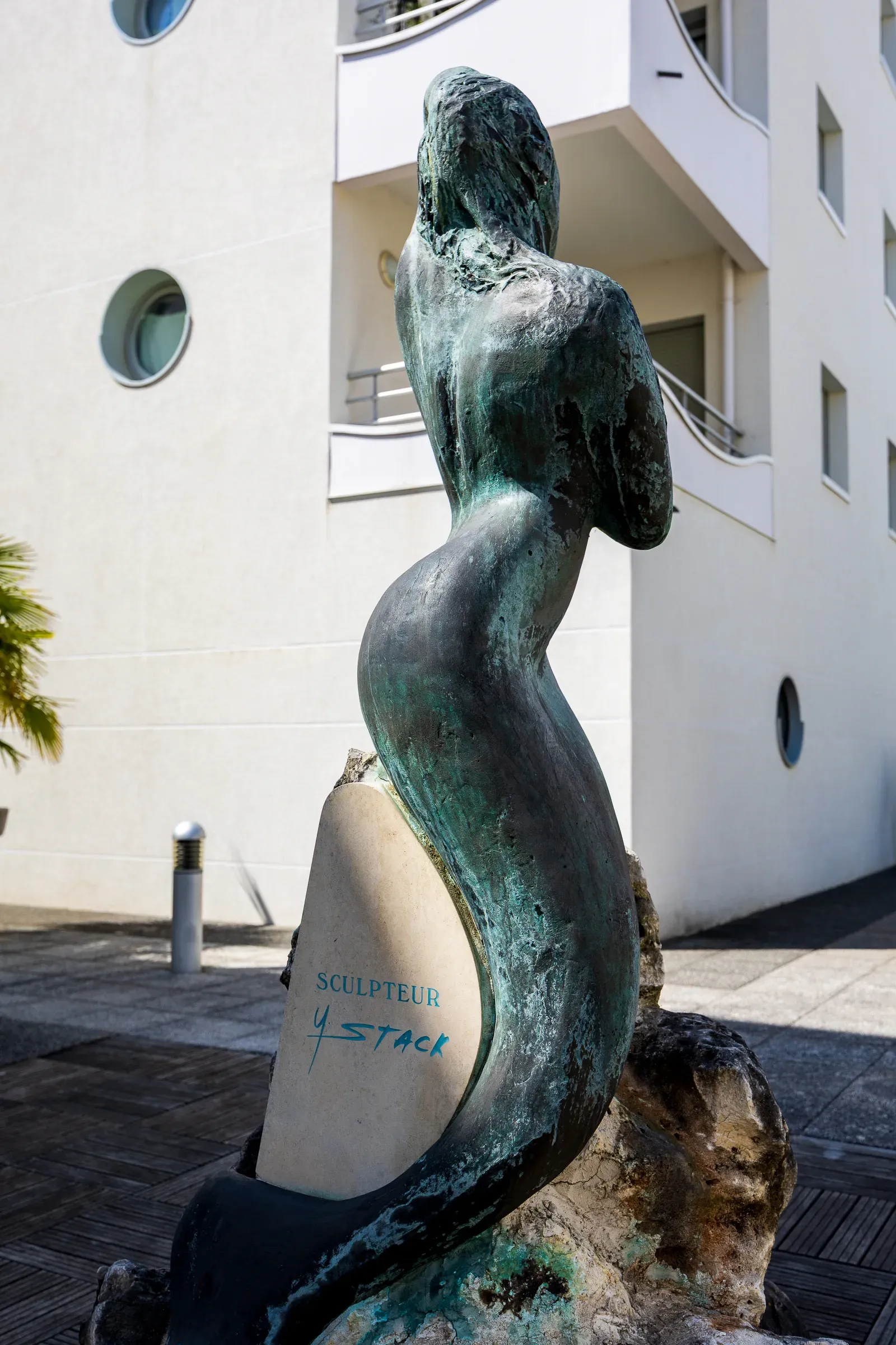 Green-patinated bronze female figure on a stone base signed 'Sculpteur Y. Stack', white modern building with circular portholes behind