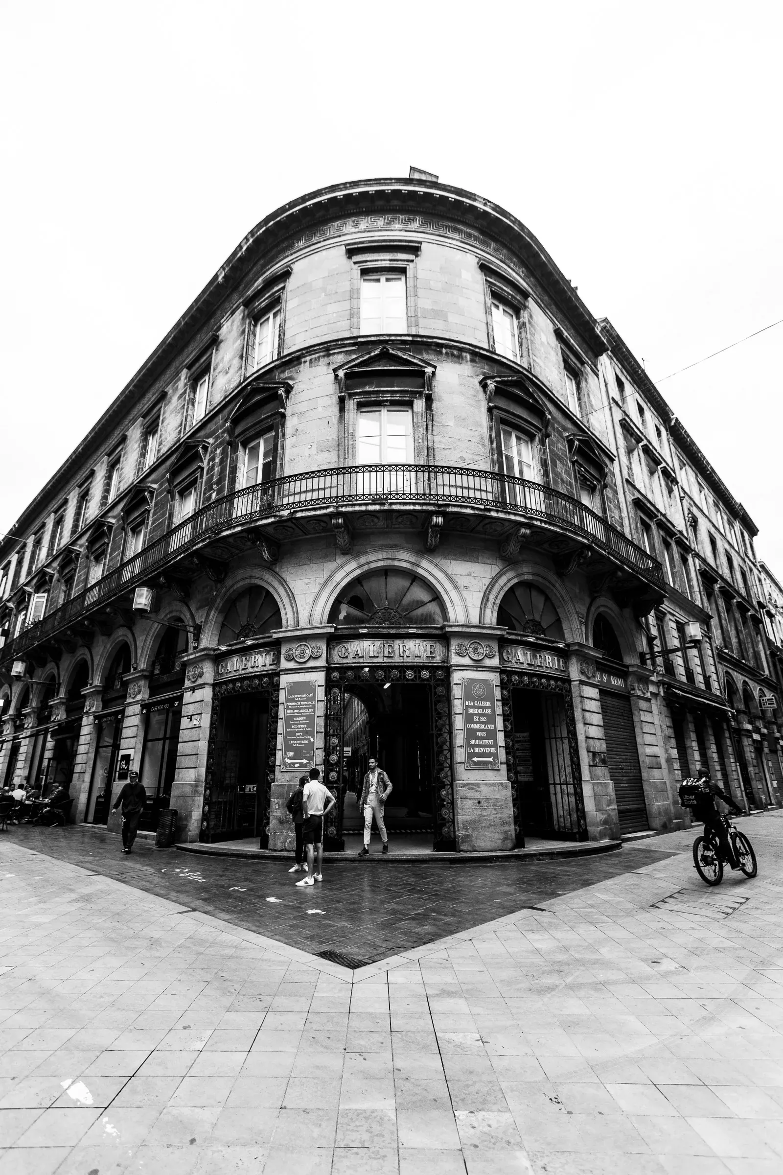 Rounded corner of a Haussmann building in Bordeaux shot with a wide angle, arched ground-floor entrances, a handful of figures on the wet pavement