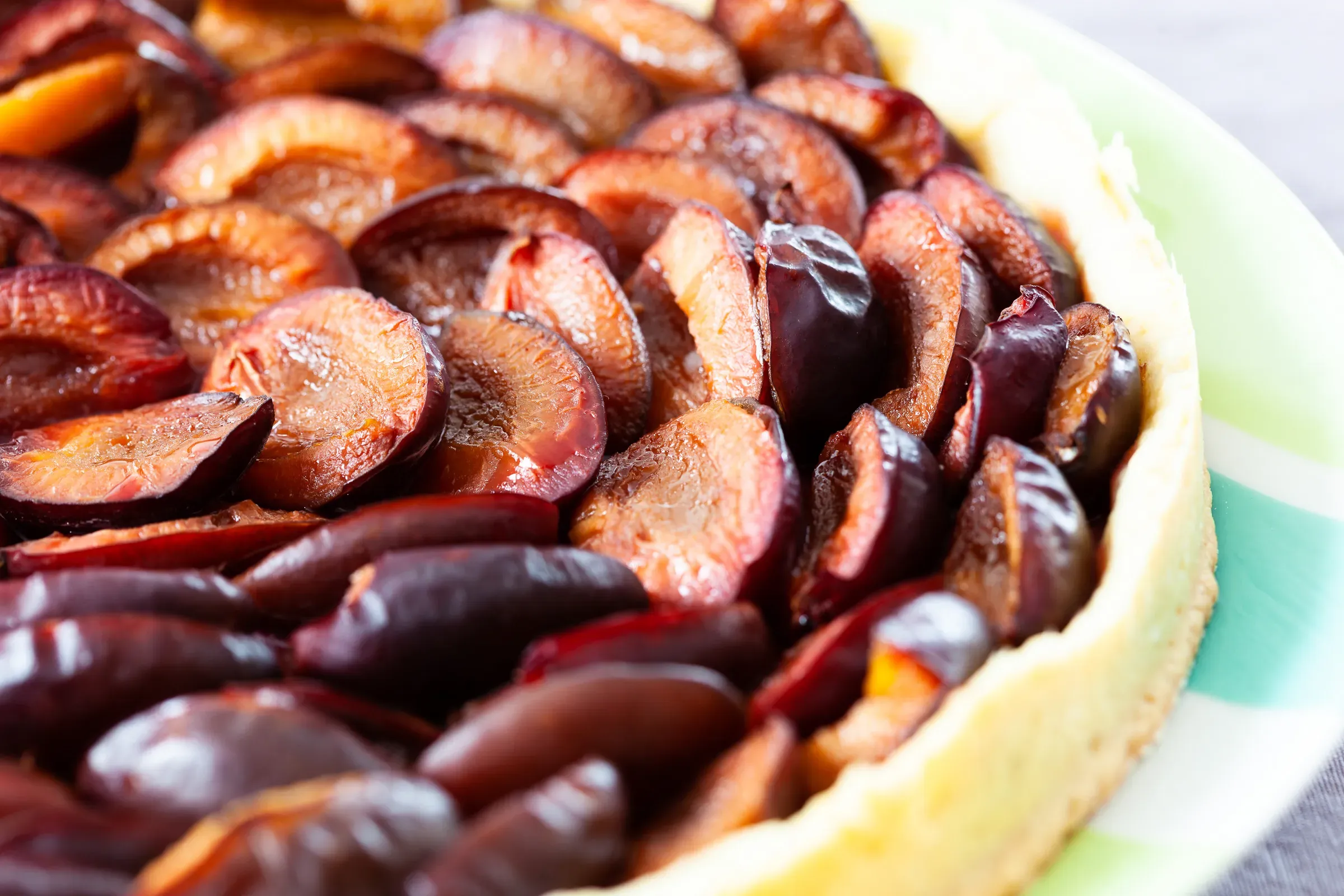 Plum tart close-up — halved prune plums packed tightly and glazed in a short pastry shell, deep purple and amber tones