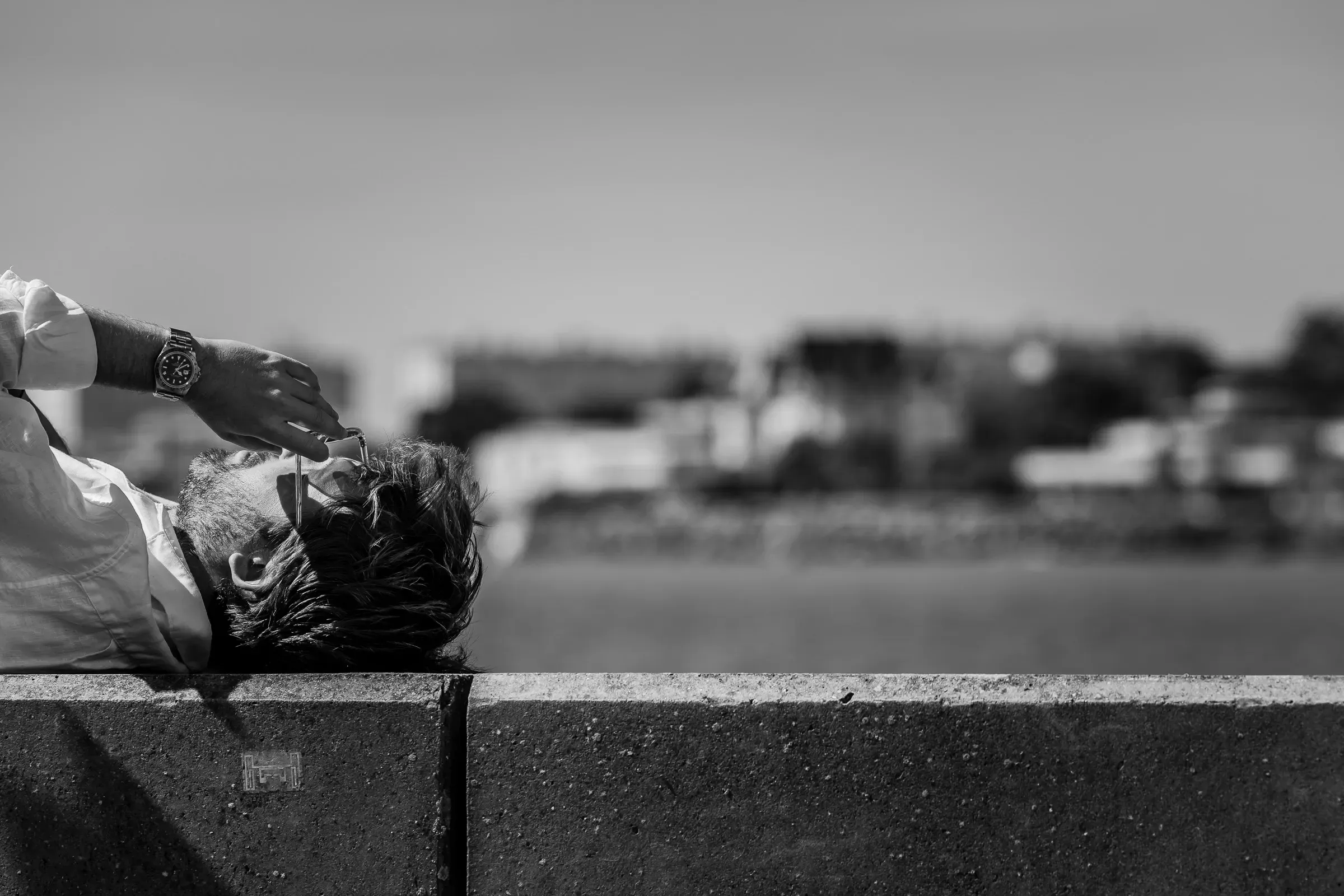 A man reclines against a wall, head tipped back, as a hand applies eye drops, a blurred skyline beyond