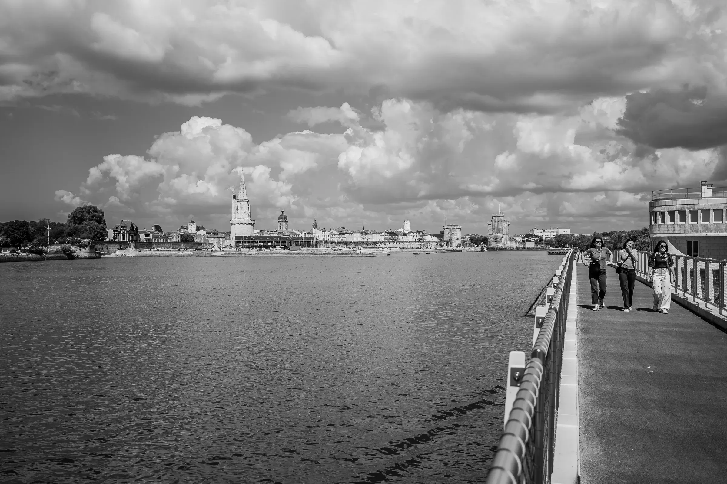 Black and white — three women walking on a waterfront bridge, La Rochelle's skyline with towers in the background, dramatic cumulus clouds overhead
