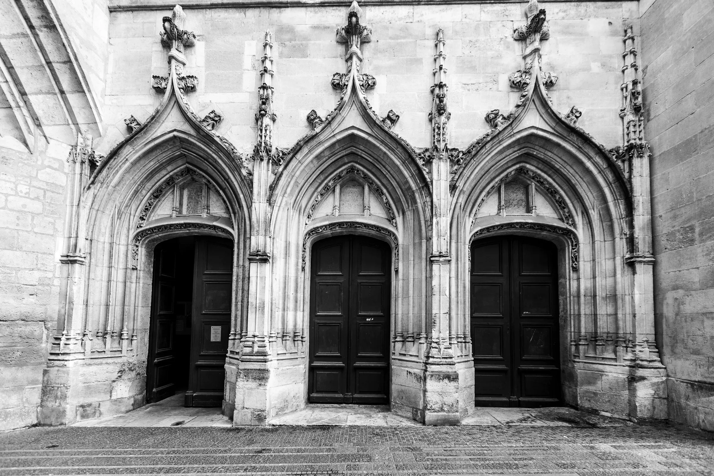 Three side-by-side Gothic arched doorways in a medieval church wall, ornate stone crockets and finials above each, heavy dark wooden doors below