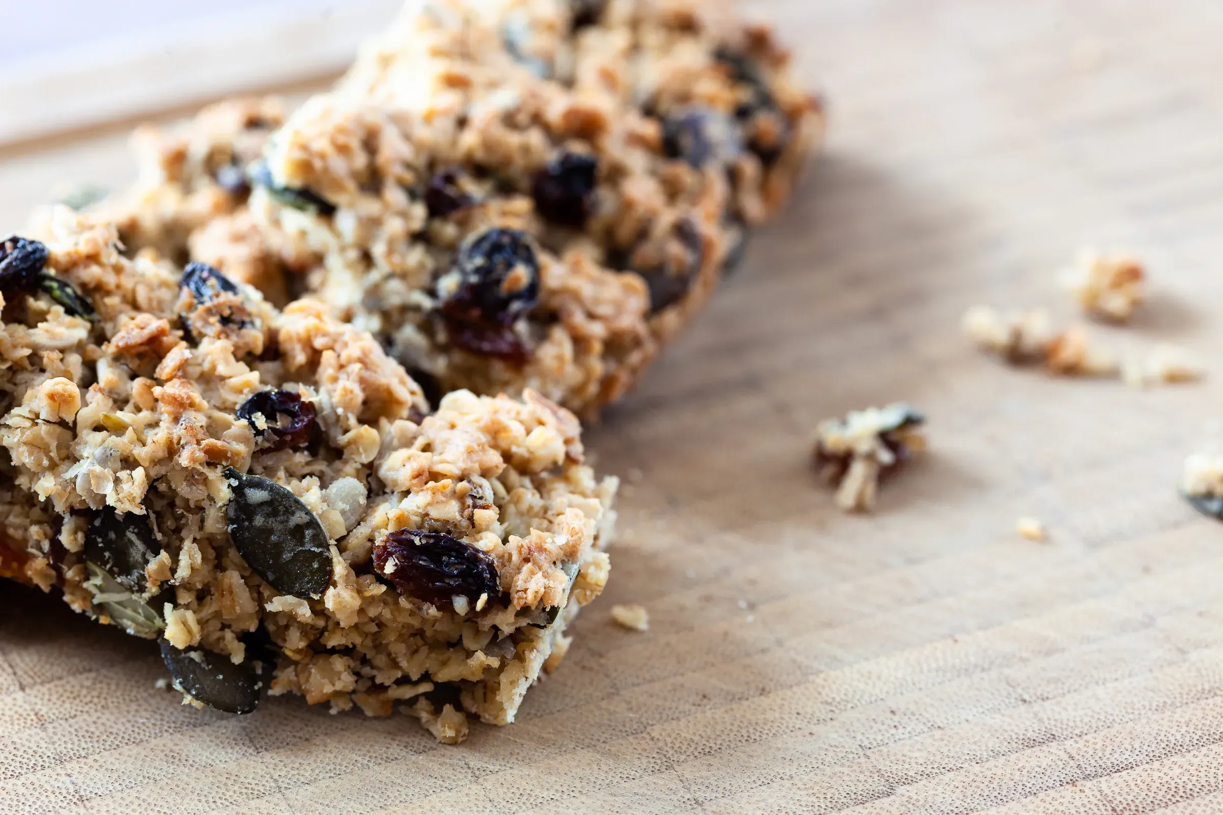 Two oat, seed and raisin flapjack bars on a wooden board, crumbs scattered around them, shot from a low angle