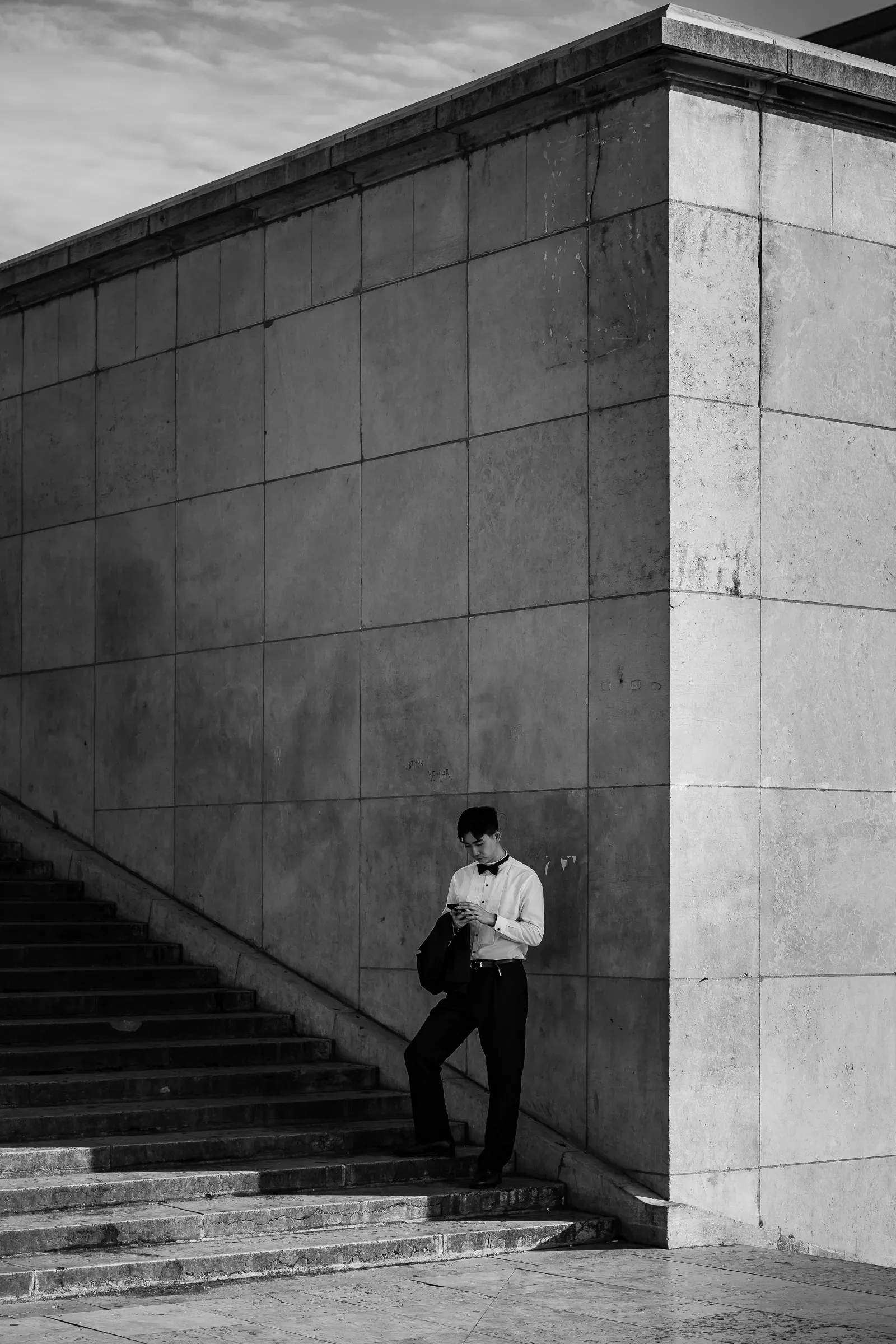 A young man in a white shirt and bow tie checks his phone on a stone staircase