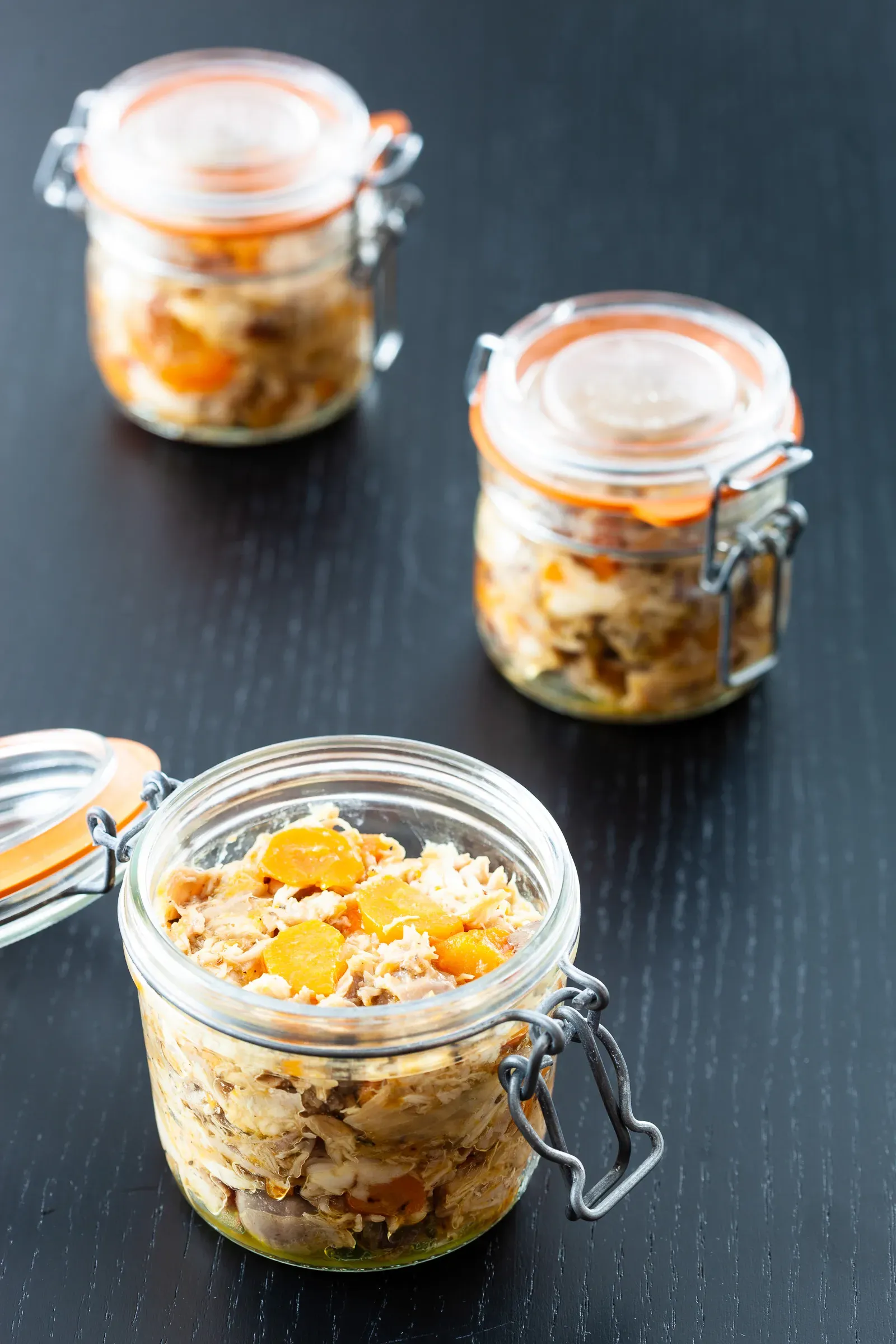 Three clip-top glass jars of rabbit rillettes on a dark wood surface — shredded meat with orange carrot pieces visible, arranged in a triangle