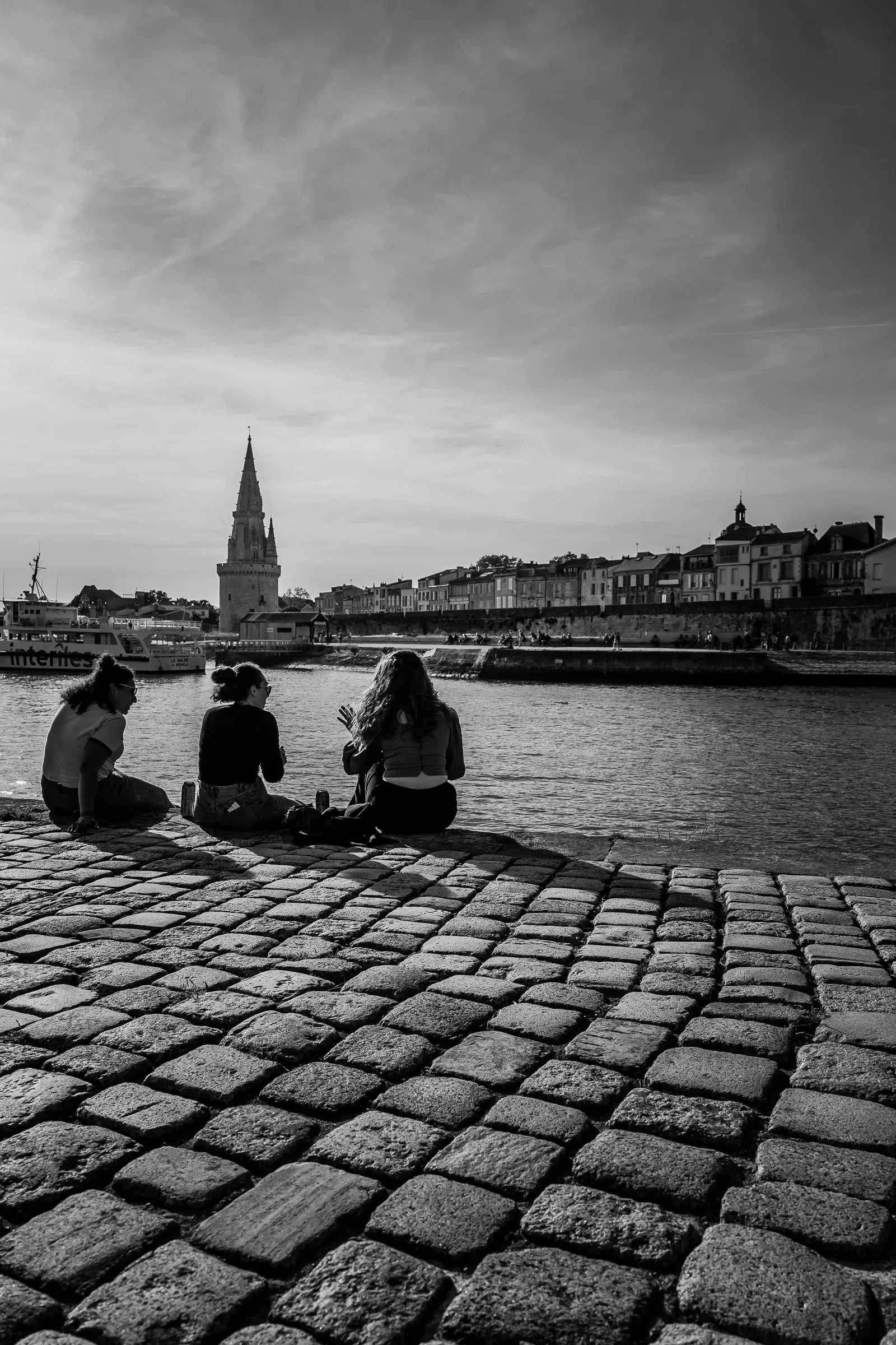 Black and white — three young women sitting on harbour cobblestones with their backs to camera, the Lanterne tower and waterfront across the water under a streaky sky