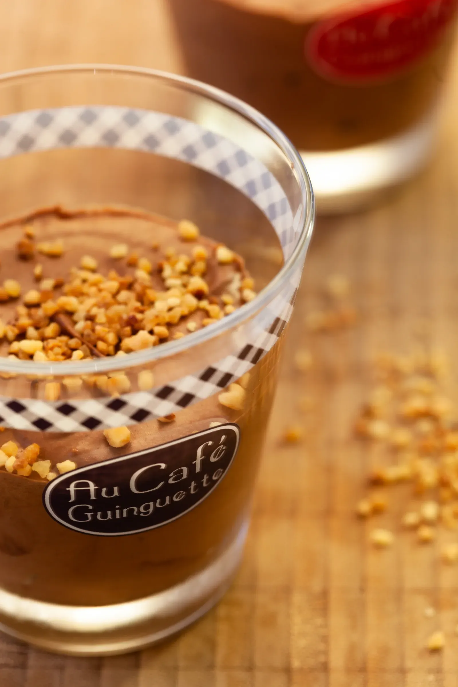 Chocolate mousse in a gingham-rimmed bistro glass labelled Au Café Guinguette, chopped hazelnuts on top, second glass and scattered nuts behind on a wooden table