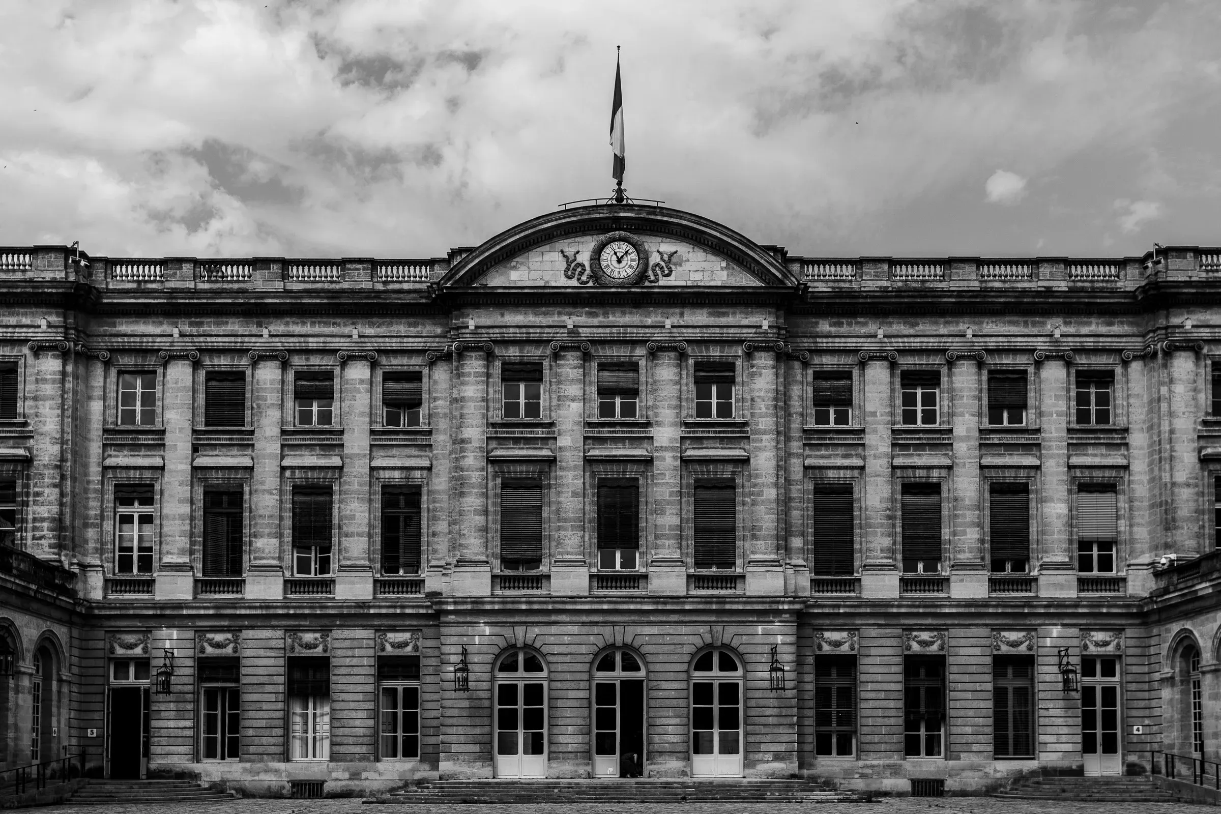 Symmetrical neoclassical facade of the Hôtel de Ville de Bordeaux, French flag at the pediment, clock above the central arch, the courtyard empty
