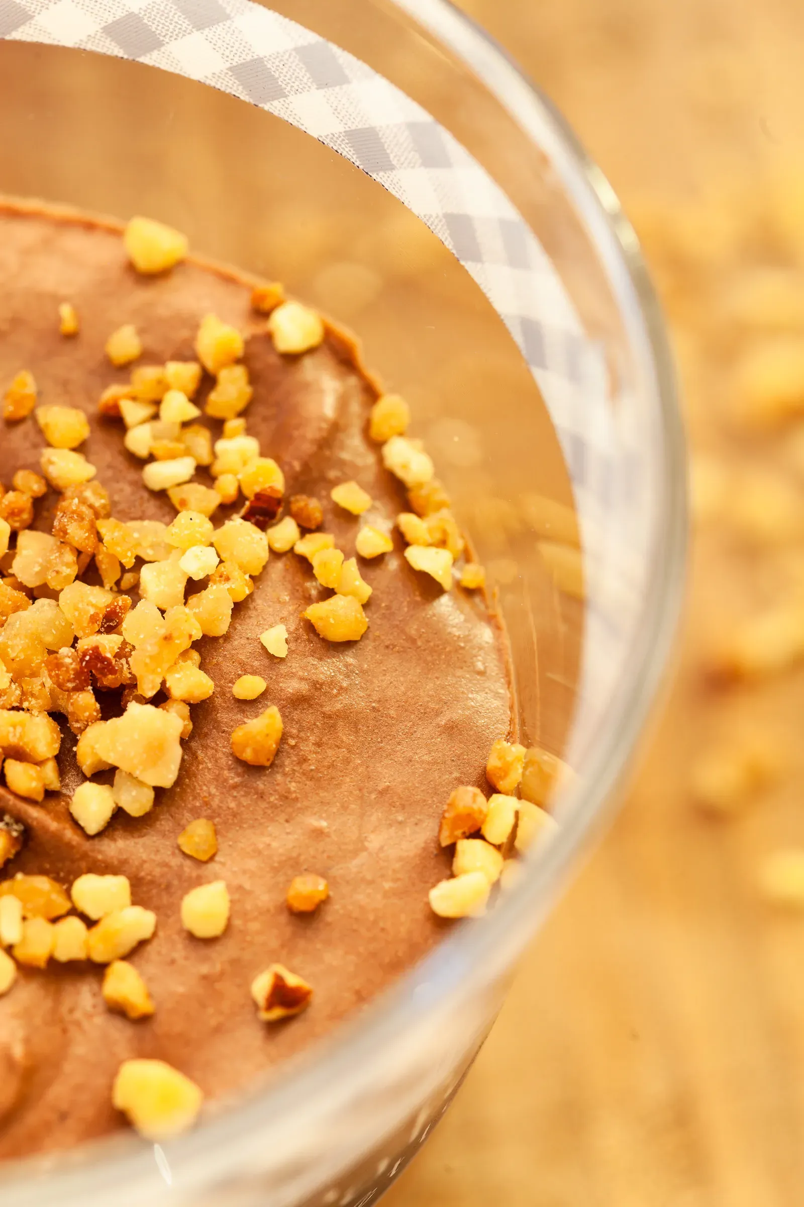 Close-up overhead of chocolate mousse surface, chopped hazelnuts scattered across the top, gingham-patterned glass rim visible