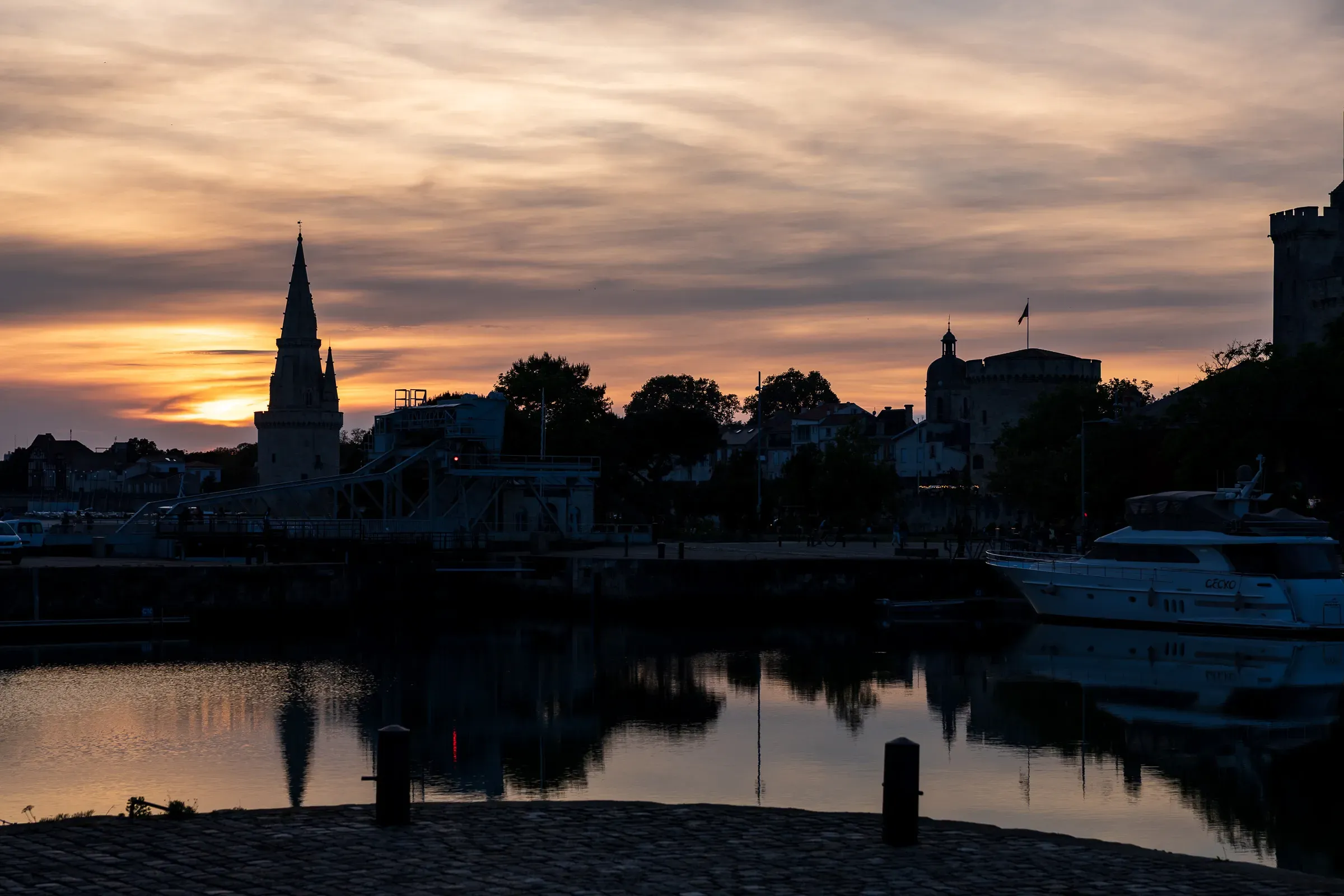 Sunset over La Rochelle's inner harbour, orange and purple sky, church spire and medieval tower silhouetted, warm reflections on dark still water