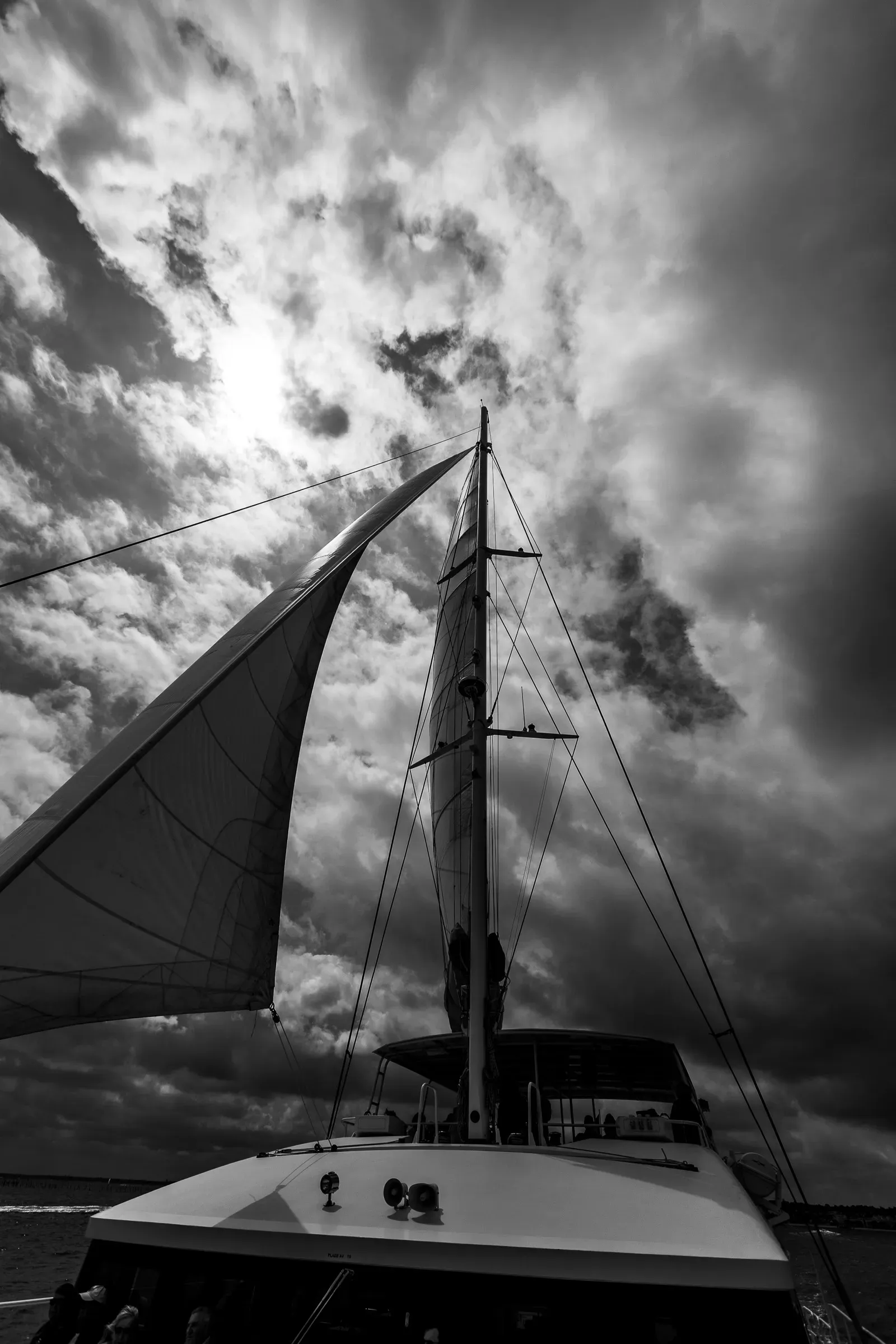 Looking straight up along a sailboat mast into a dramatic cloudy sky, the mainsail taut on the left, rigging lines cutting across the frame