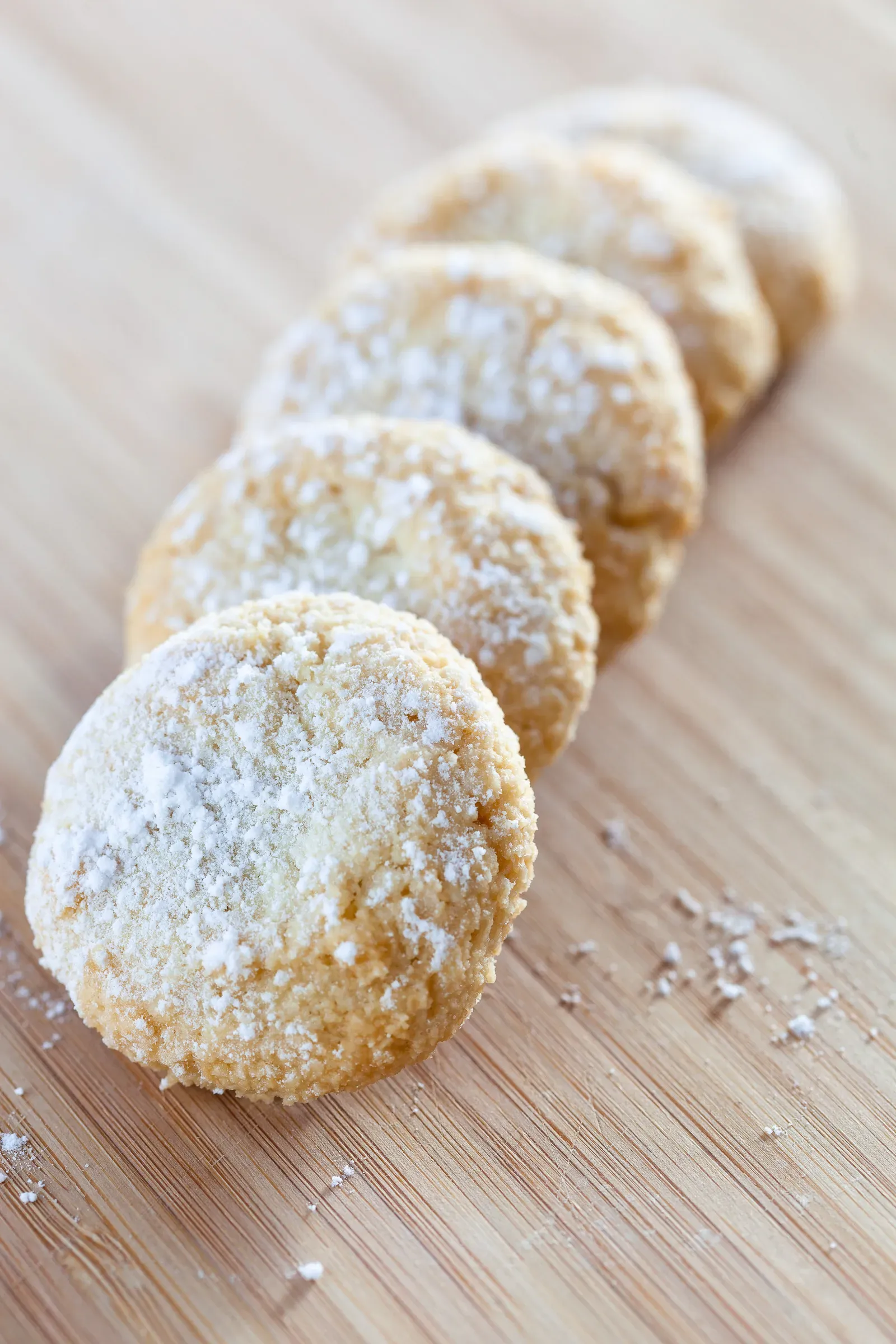 Five sugared shortbread rounds lined up in a row on a wooden surface, powdered sugar dusted, receding into soft focus