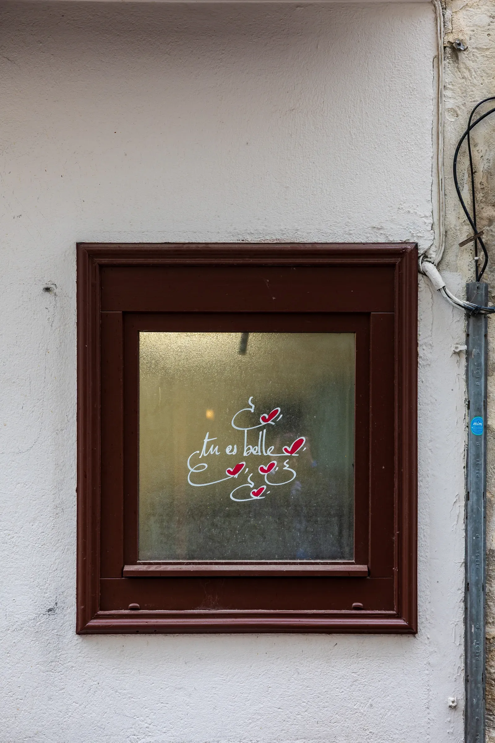 Small dark-framed window in a whitewashed wall with 'tu es belle' written in cursive on the glass and small red heart decorations