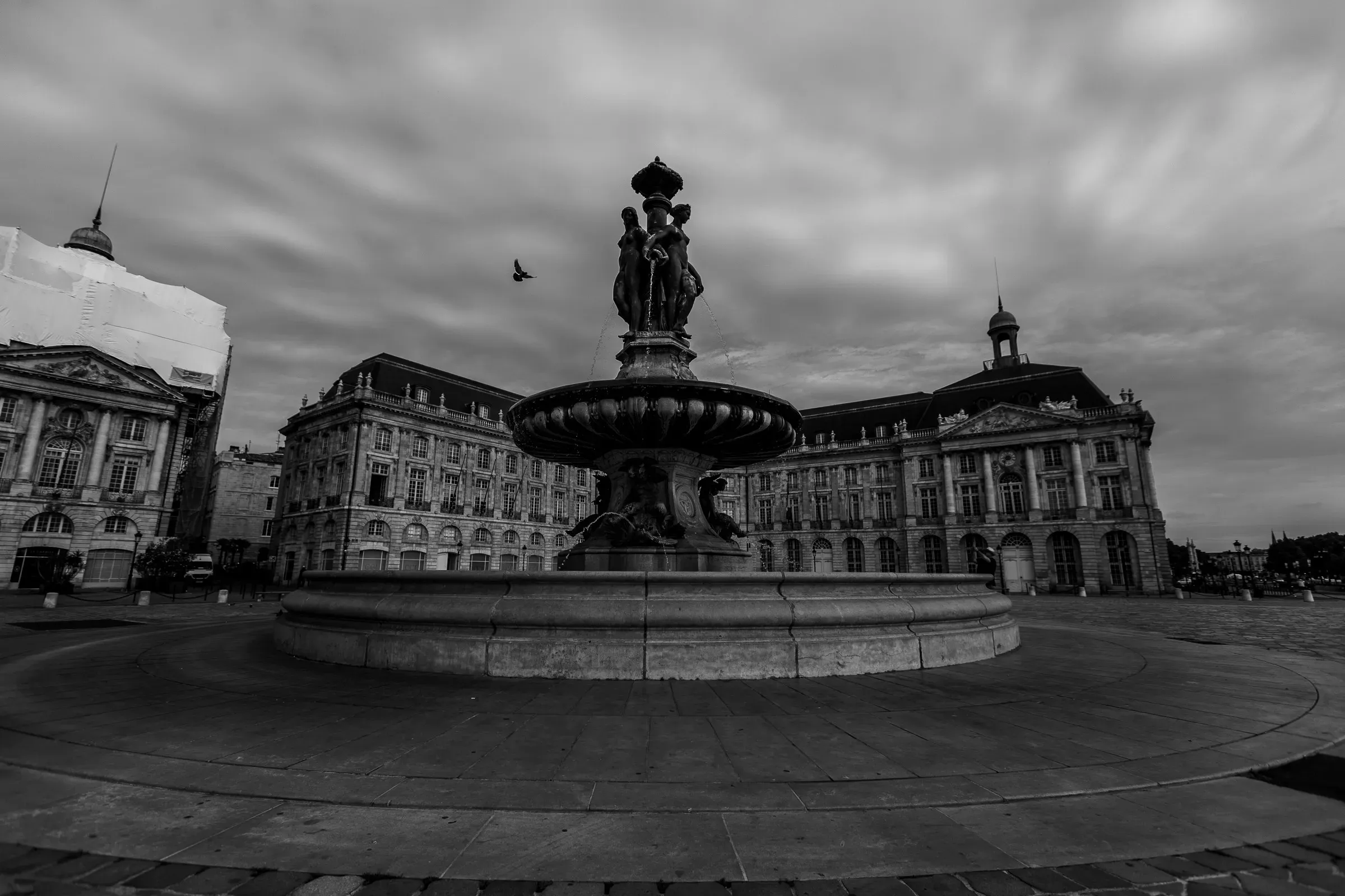 Fountain of the Three Graces on Place de la Bourse in Bordeaux, wide angle, a single bird crossing the moody long-exposure sky, the Bourse behind