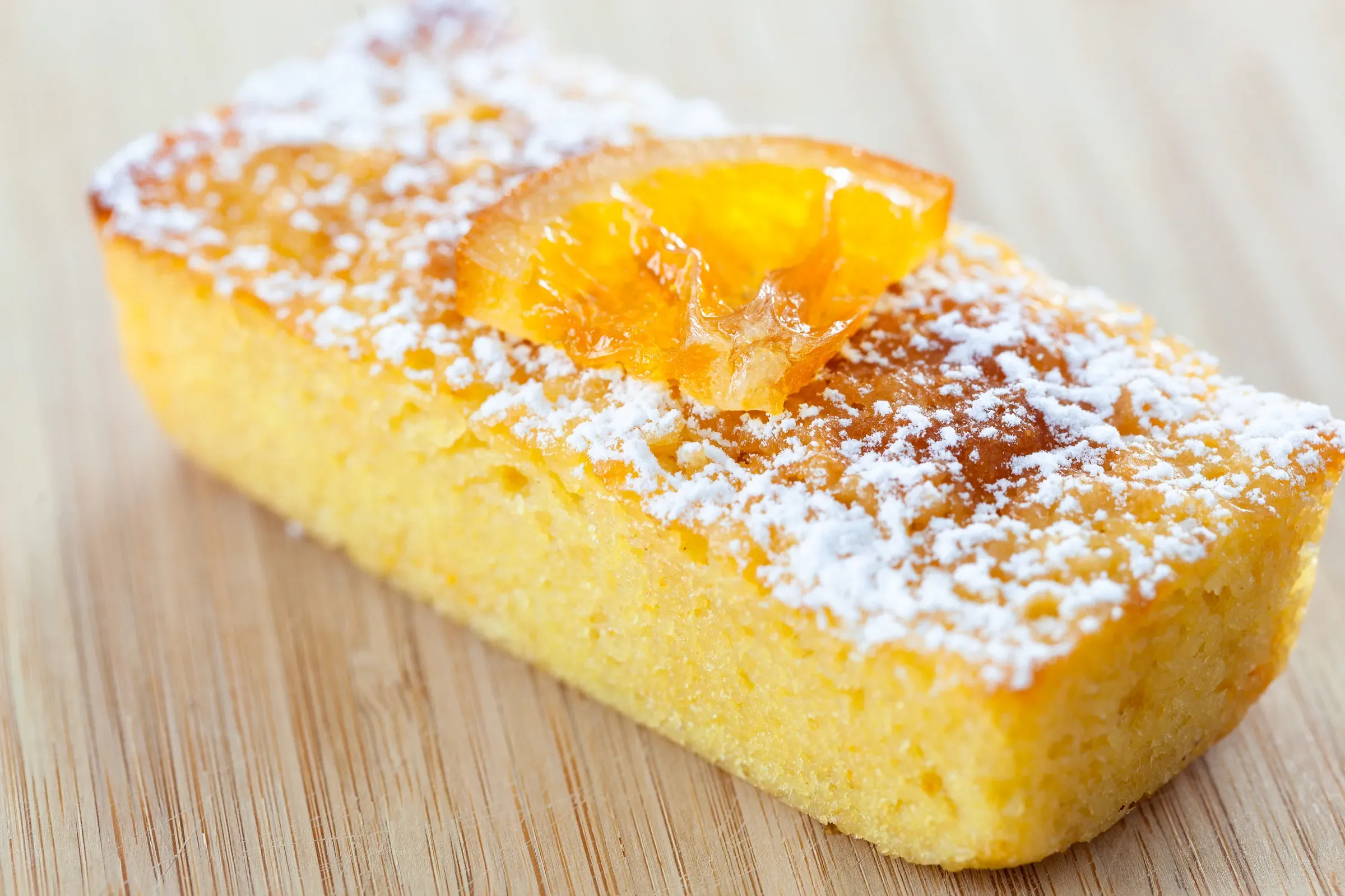 Orange loaf cake on a wooden board, a candied orange slice on top, dusted with powdered sugar, pale crumb visible at the cut end