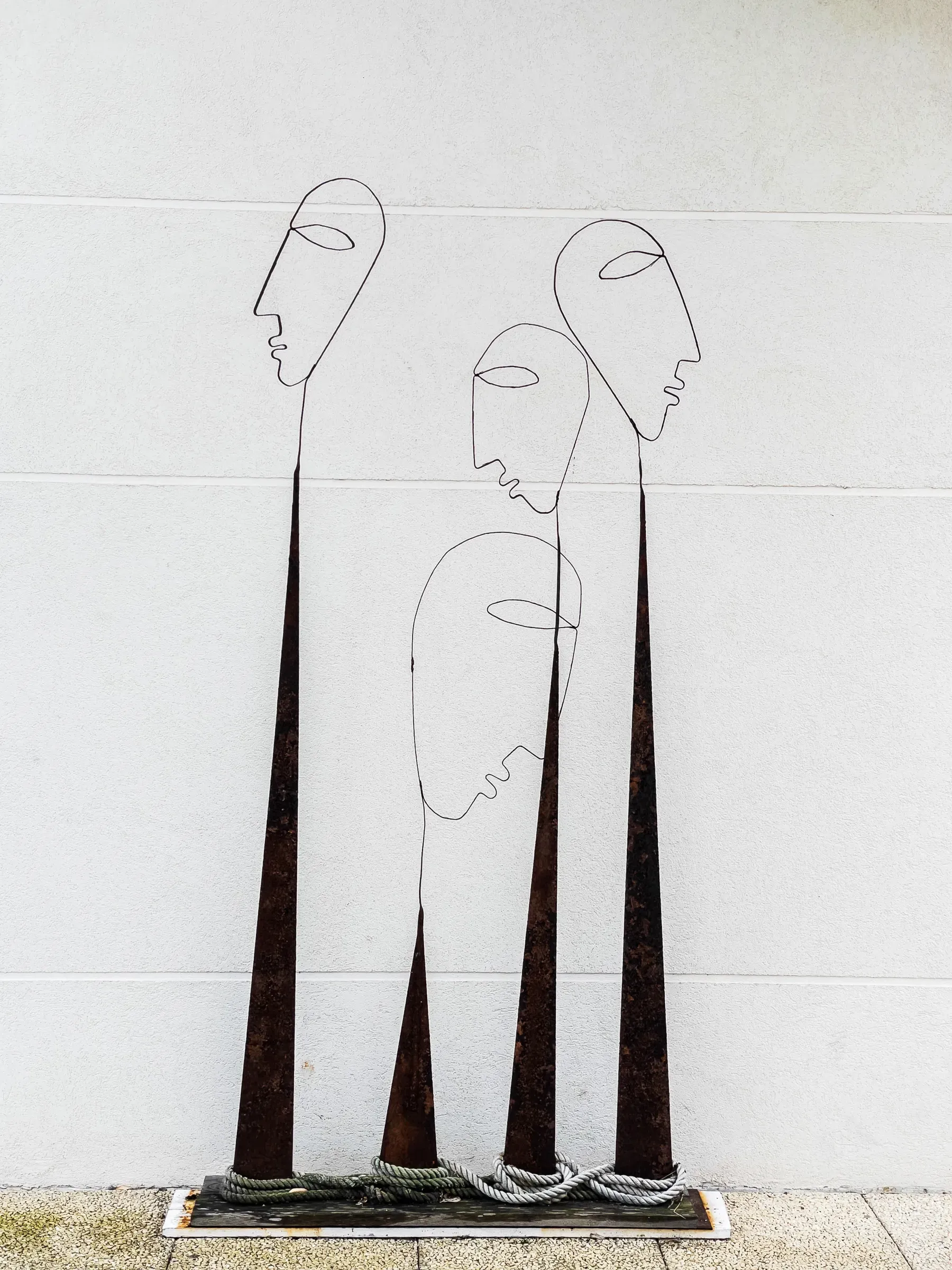 Three elongated wire figures with blank oval faces on rusted metal bases, rope coils at their feet, white wall behind