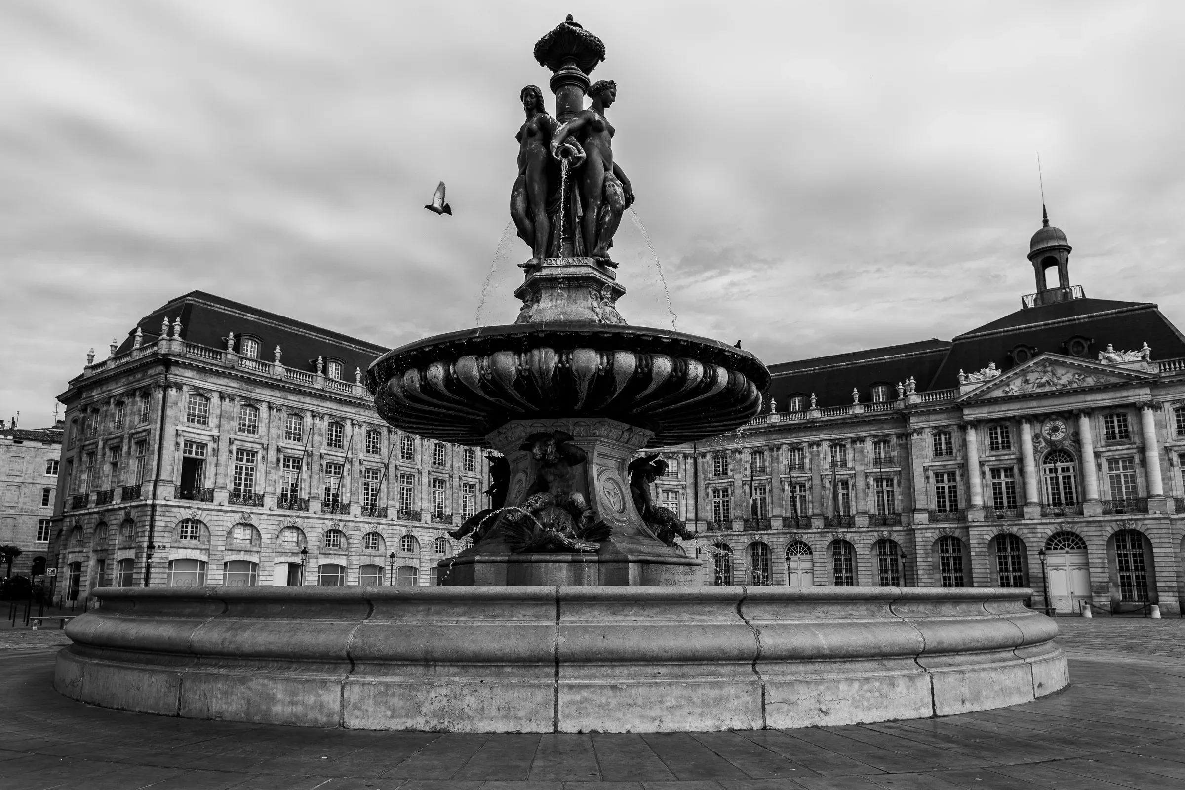 Fountain of the Three Graces at closer range, the sculpted figures and basin sharp, the Bourse building across the square, a bird suspended mid-flight