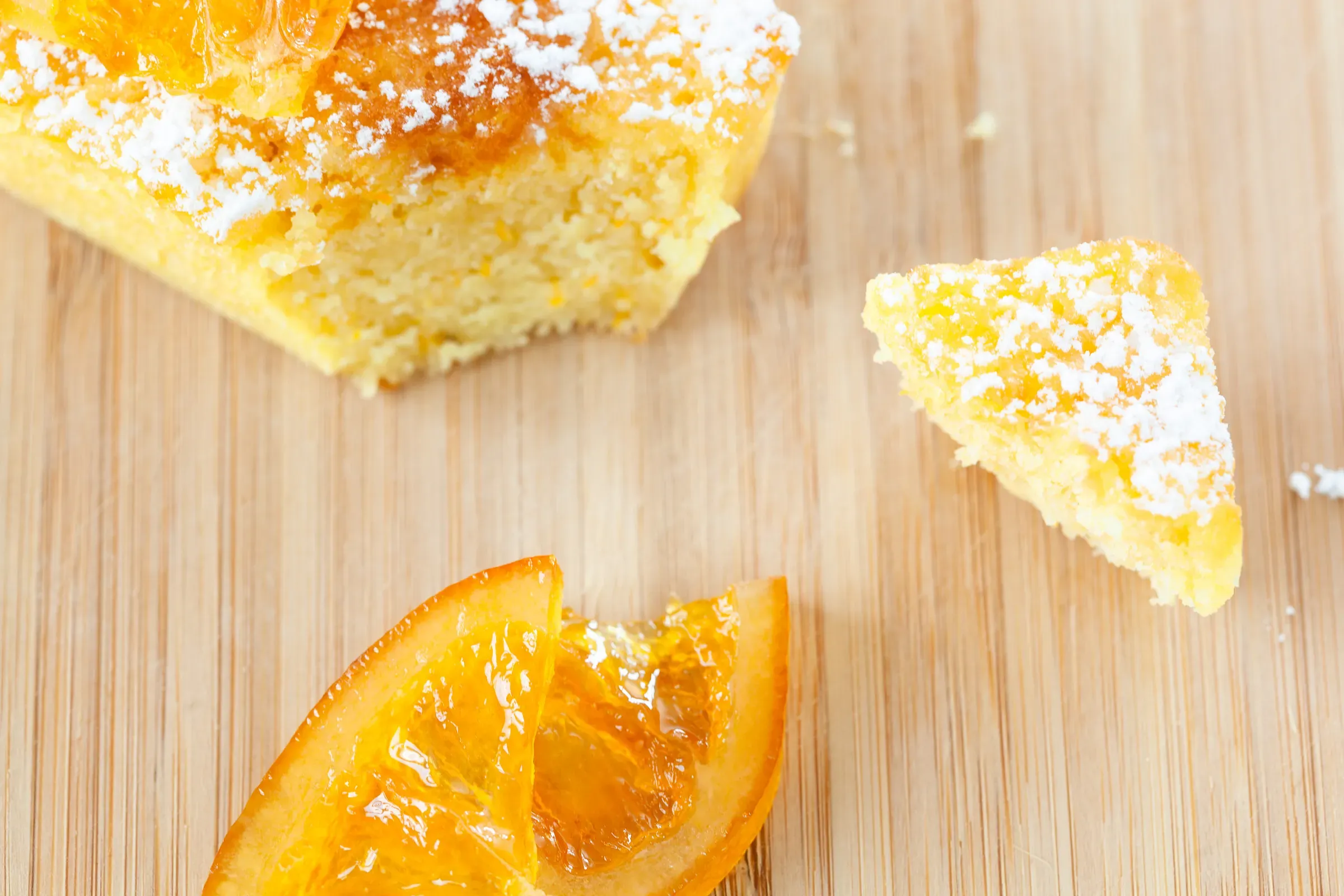 Orange cake overhead on a wooden board — a cut slice beside the loaf, two candied orange wedges, powdered sugar dusting