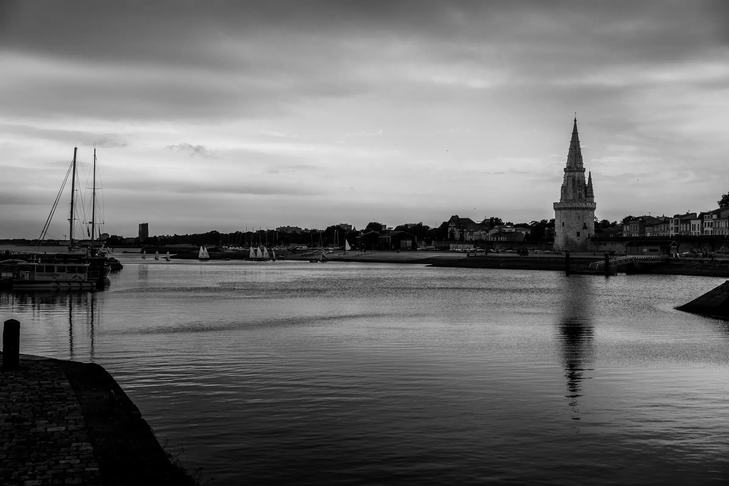 Black and white — La Rochelle harbour at dusk, the Lanterne tower spire reflected in calm dark water, sailboat masts to the left, overcast sky above