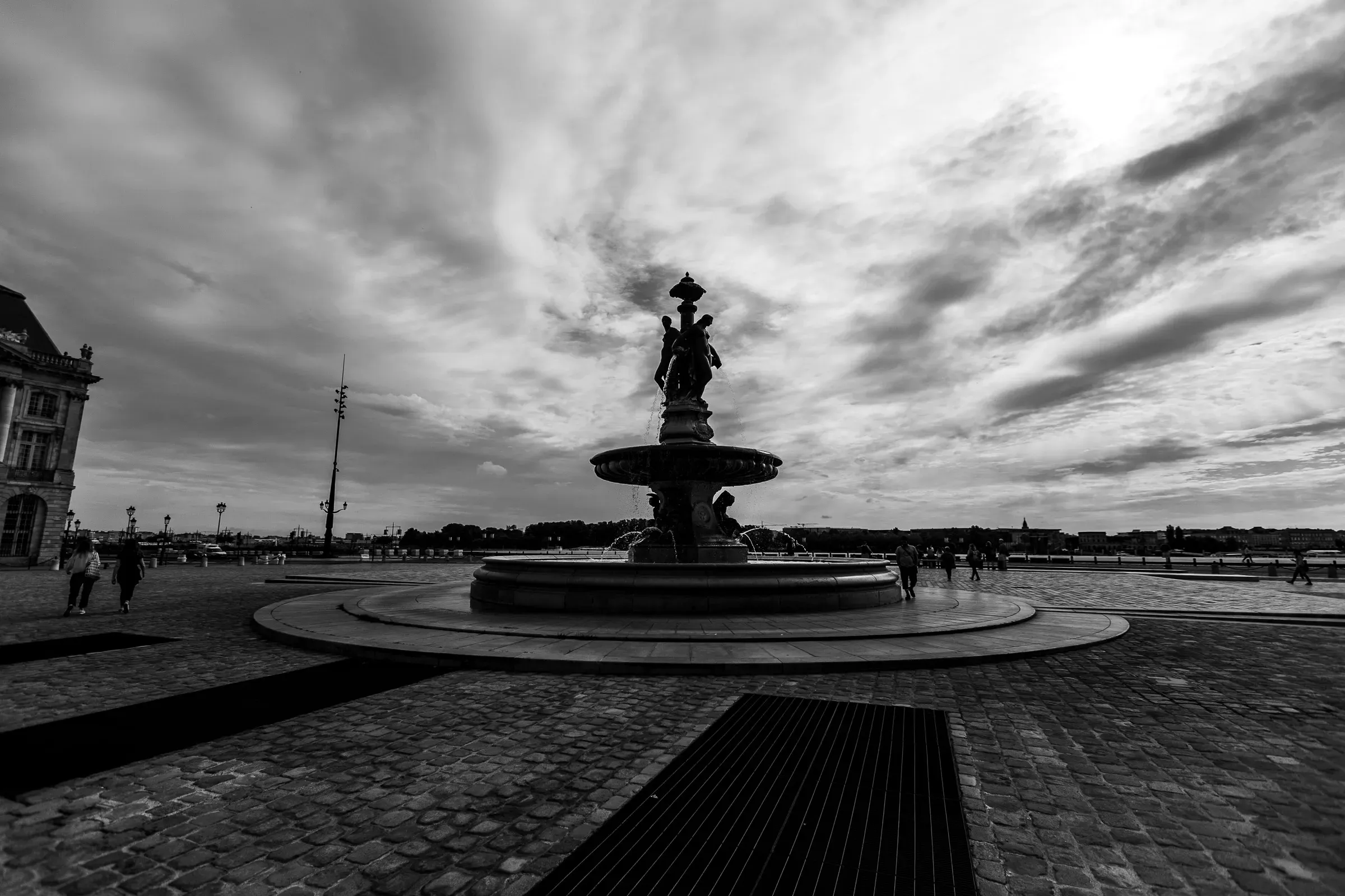 Three Graces fountain as a dark silhouette against a wide dramatic sky, the Garonne and a bridge stretching behind, passersby in shadow