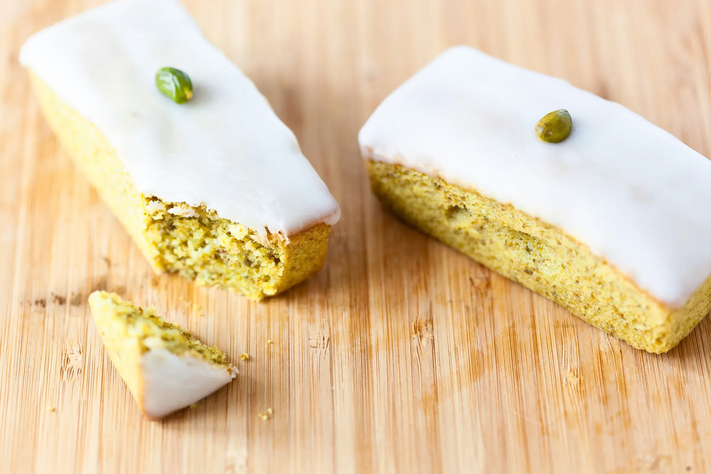 Two small pistachio loaf cakes with white glaze and a single pistachio on top each, one sliced to show the green crumb, wooden board