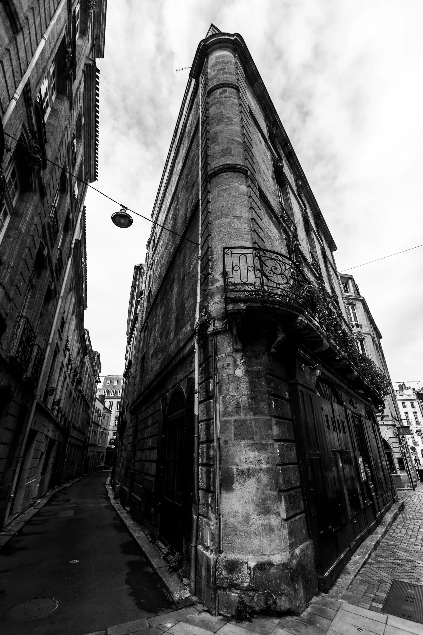 Narrow medieval street in Bordeaux's Saint-Pierre quarter, a tall rounded stone corner building, wrought-iron balcony, a pendant lamp above the lane