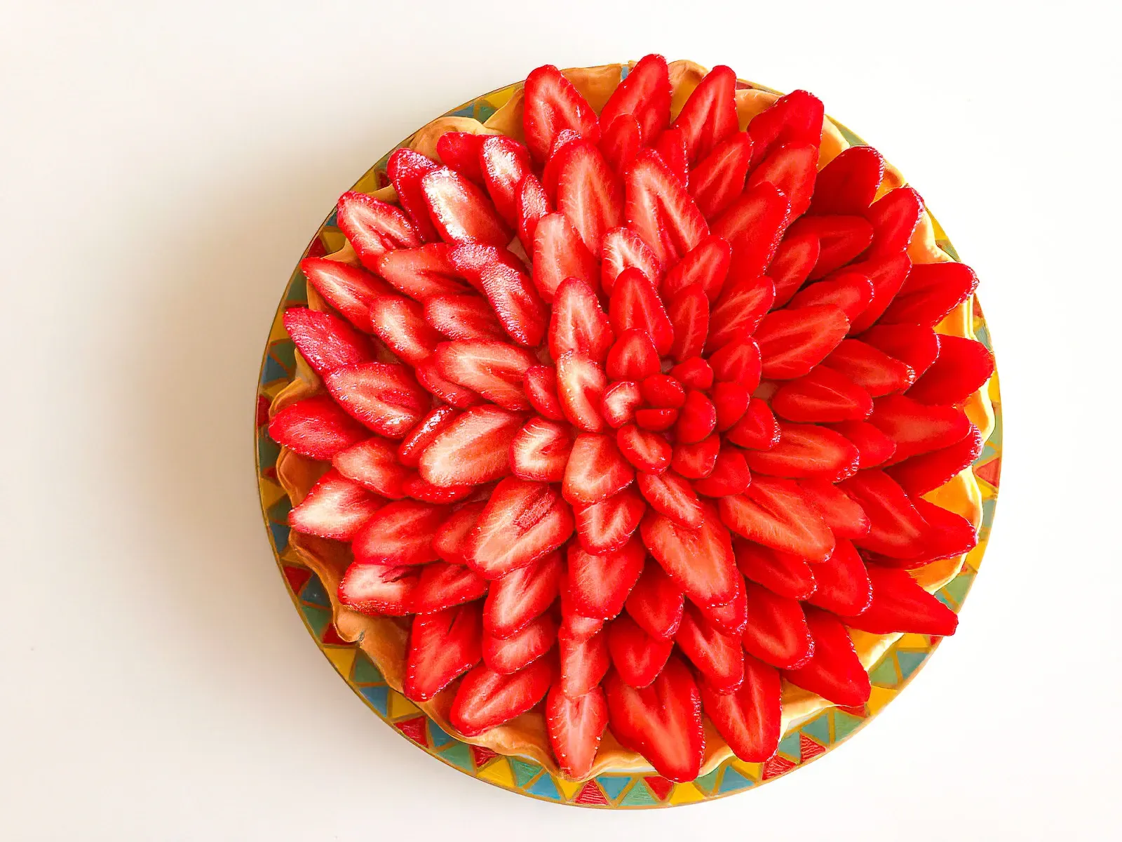 Strawberry tart overhead — strawberry slices arranged in a tight outward spiral covering the entire surface, on a colourful patterned plate, white background