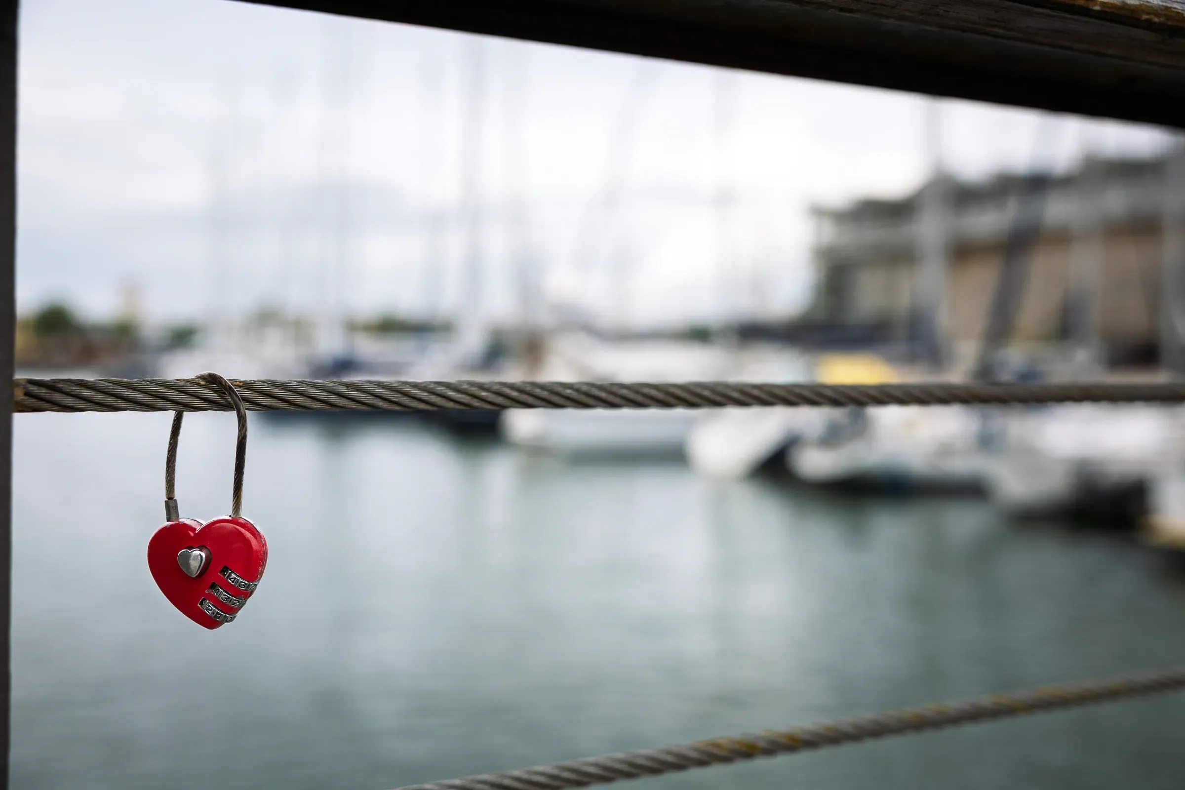 Red heart-shaped padlock clipped to a steel wire railing, harbour and moored sailboats softly blurred behind
