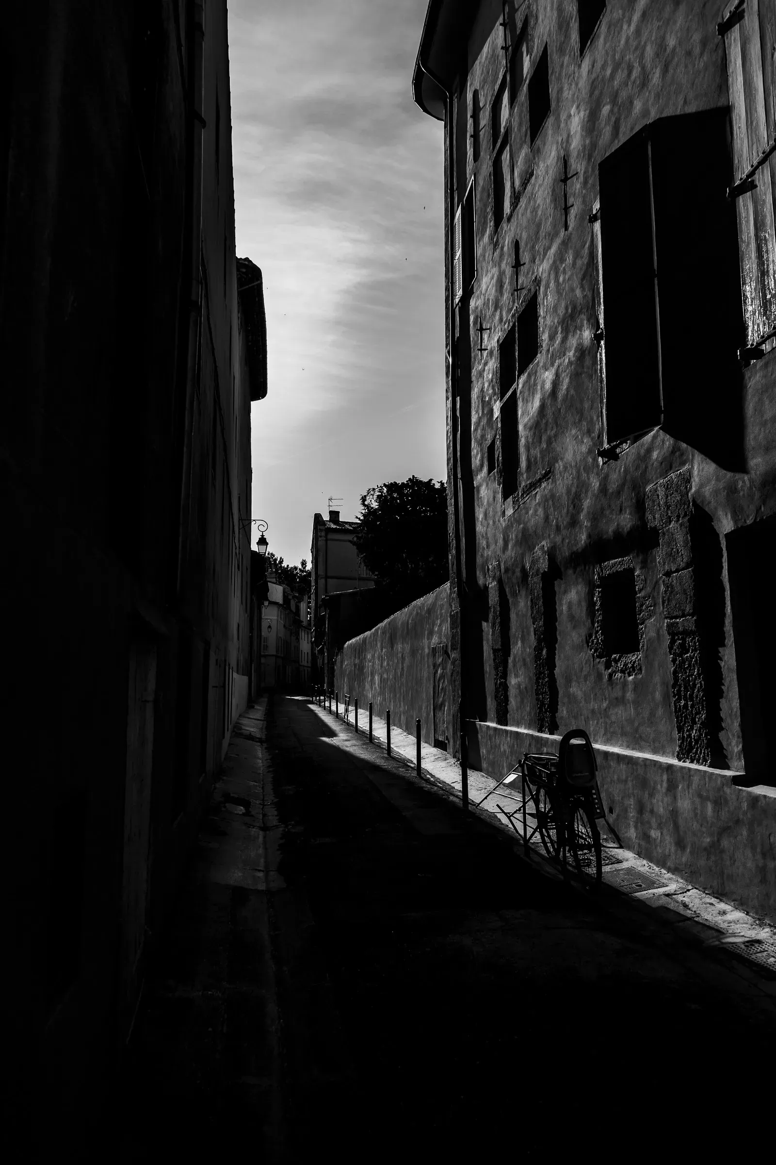 Dark narrow alley in a French town, buildings in deep shadow on either side, a solitary chair catching the light at the far end