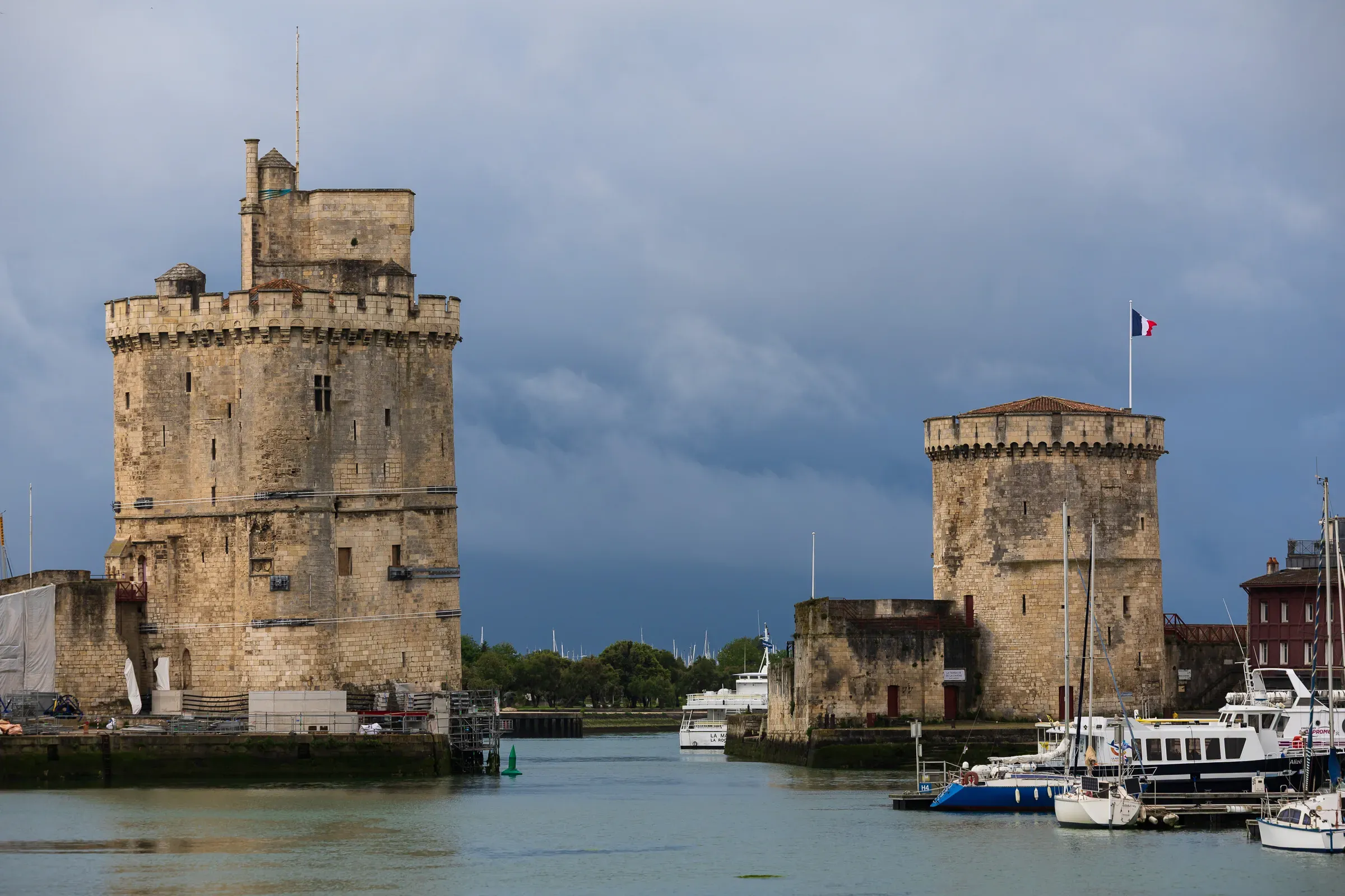 Tour Saint-Nicolas and Tour de la Chaîne under a stormy dark sky, French flag flying from the smaller tower, boats moored in the harbour entrance