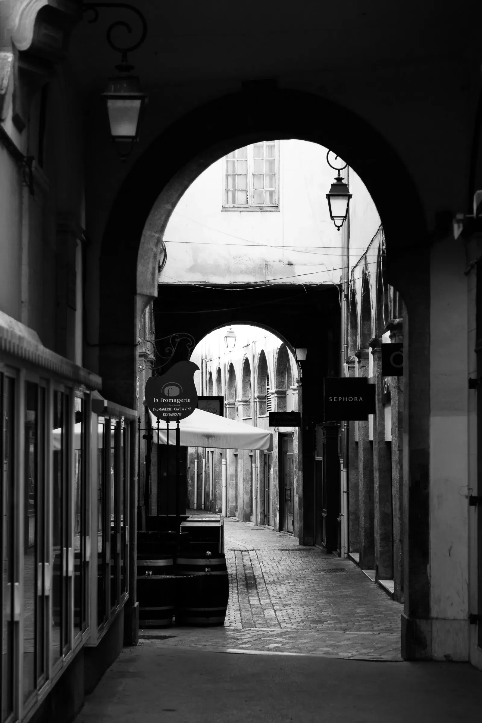 Covered arcade in a French city, successive arches receding into the distance, a cheese shop and Sephora sign visible, tables on the left