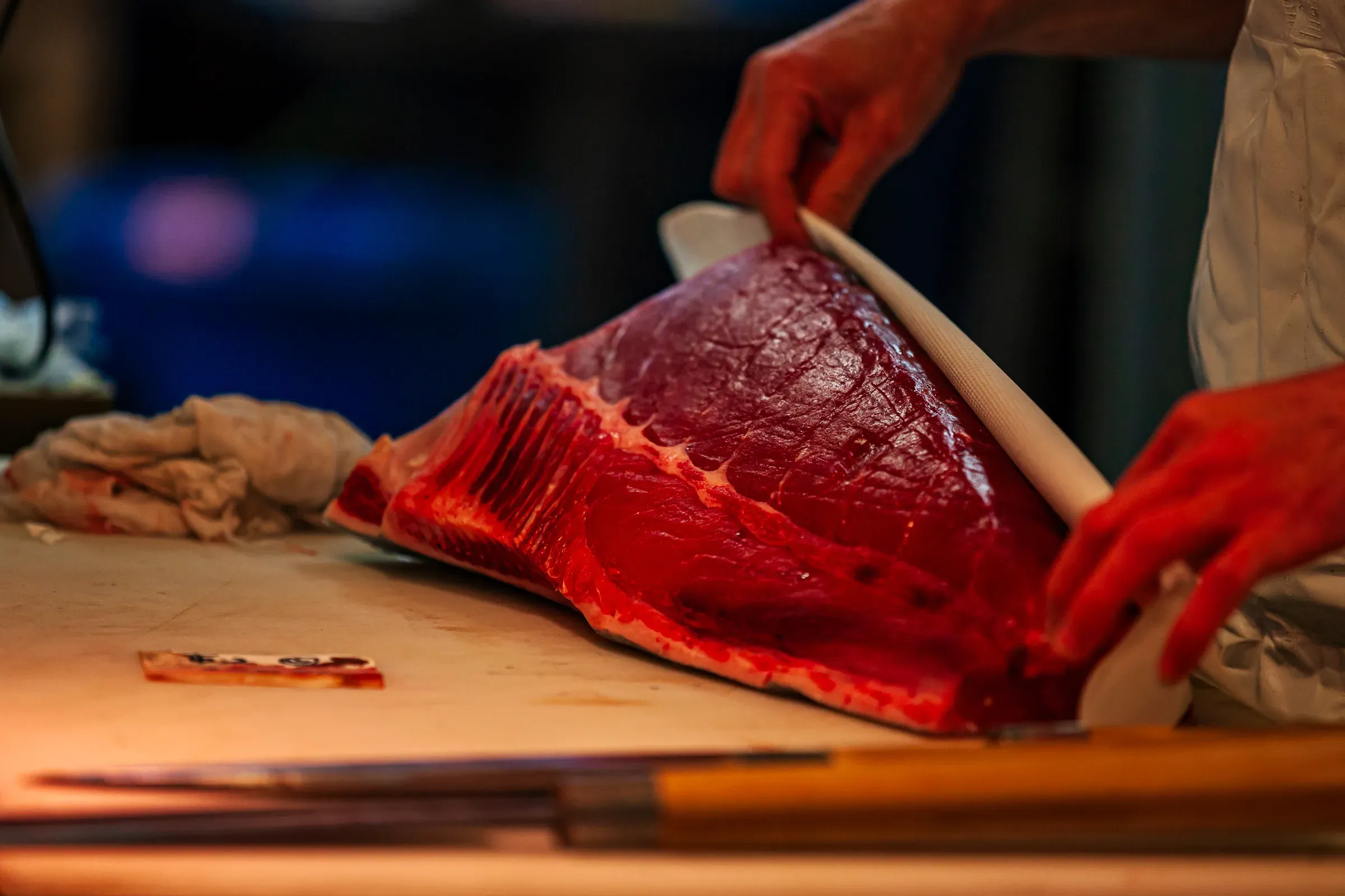 Chef's hands trimming a large loin of bluefin tuna on a cutting board under warm ambient light, blade and cloth visible