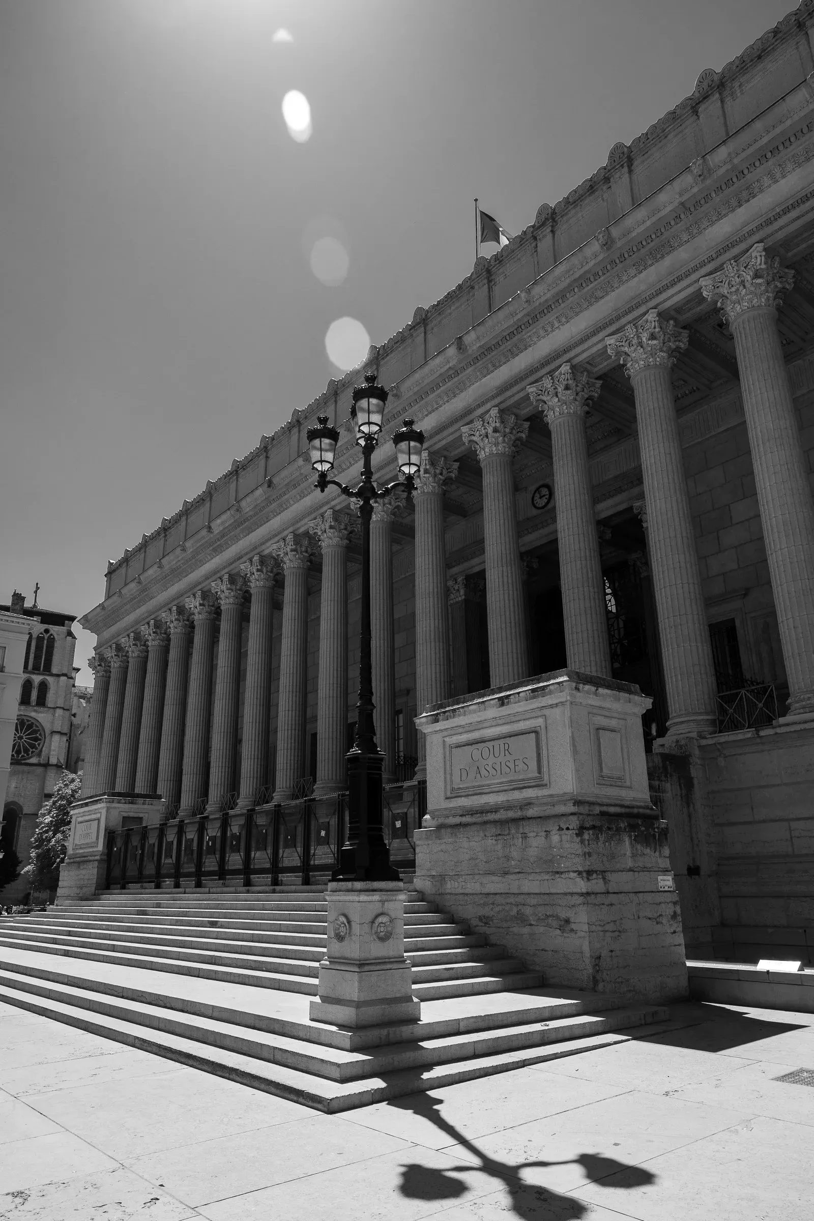 Neoclassical courthouse with a colonnade of Corinthian columns and wide stone steps, a double-headed lamp post in front, a lens flare in the bright sky