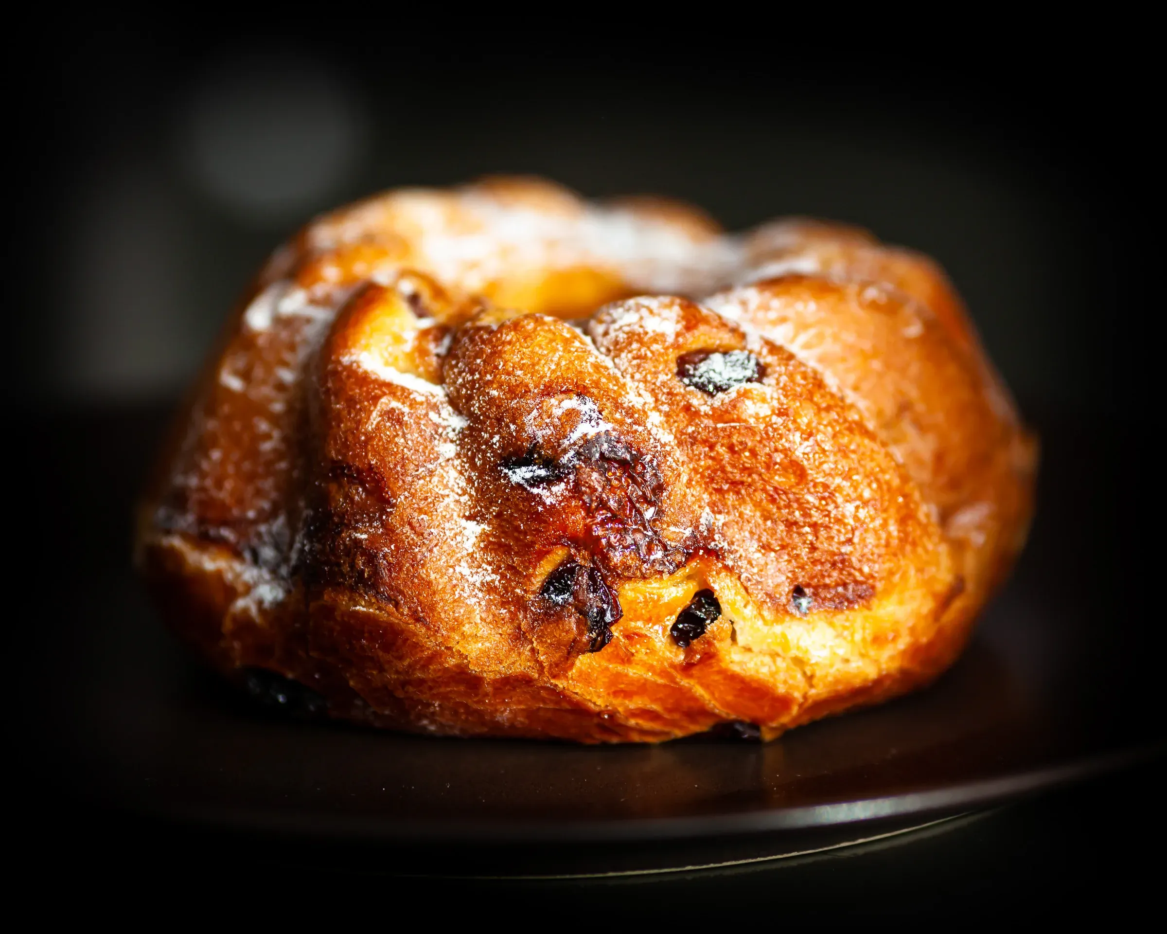 Kouglof on a dark plate against a black background — deep golden crust, raisins visible across the surface, dramatic side lighting